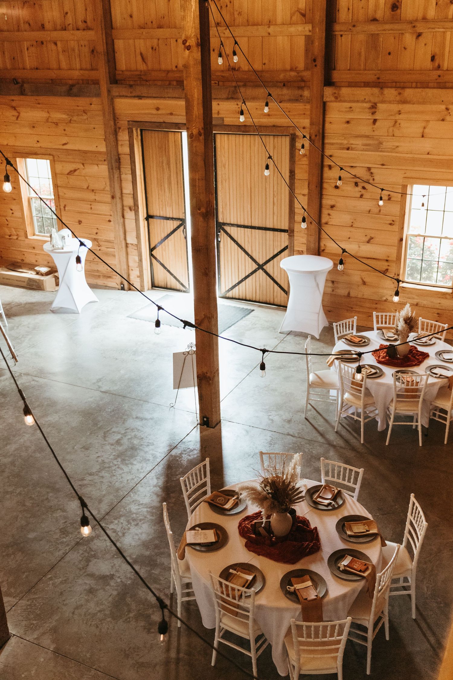 Modern rustic wedding venue interior with white chairs, string lights, and wooden beams in an elegant barn setting.