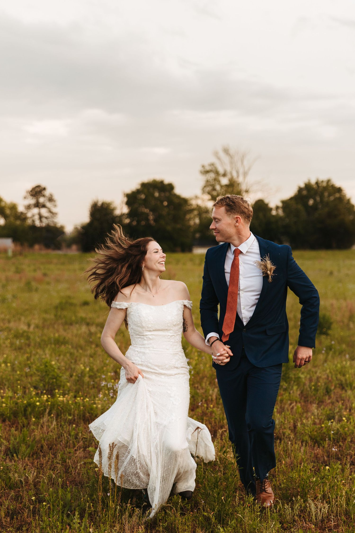 Newlywed couple runs playfully through a wildflower meadow at sunset wearing navy suit and white wedding dress.