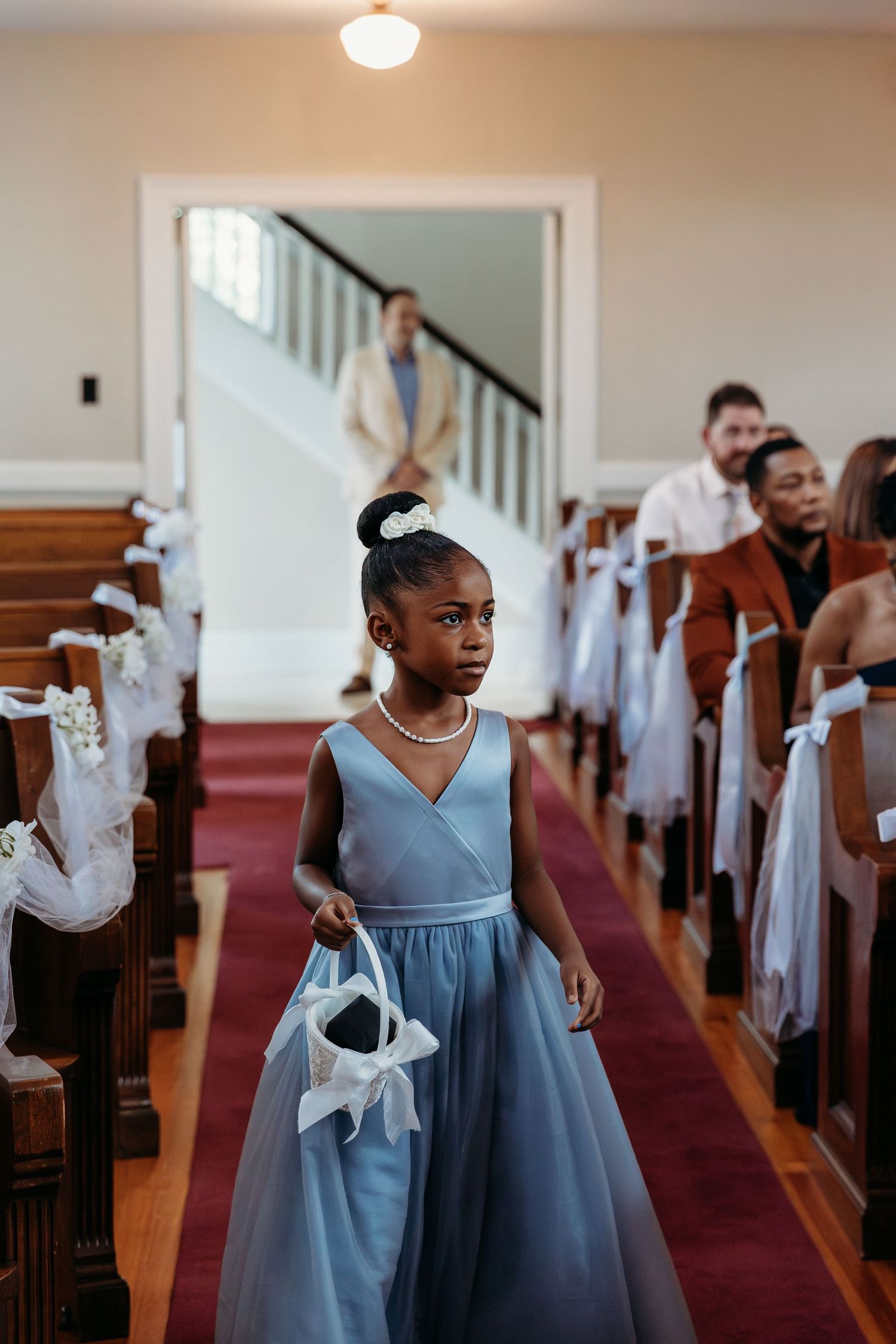 A church aisle decorated with white bows on pews shows a young attendant in a light blue dress walking down the red carpet.