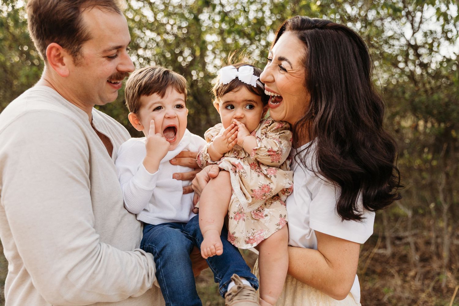 A happy family wearing coordinated white and floral outfits embraces and laughs together outdoors during a photo session.