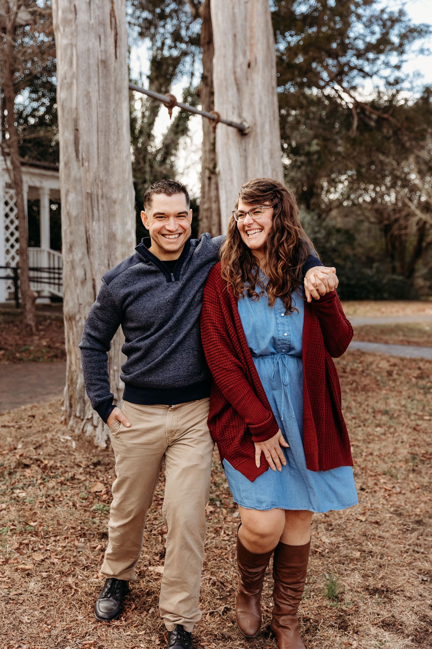 A couple in casual fall outfits laughing and posing together on fallen leaves in an outdoor setting.