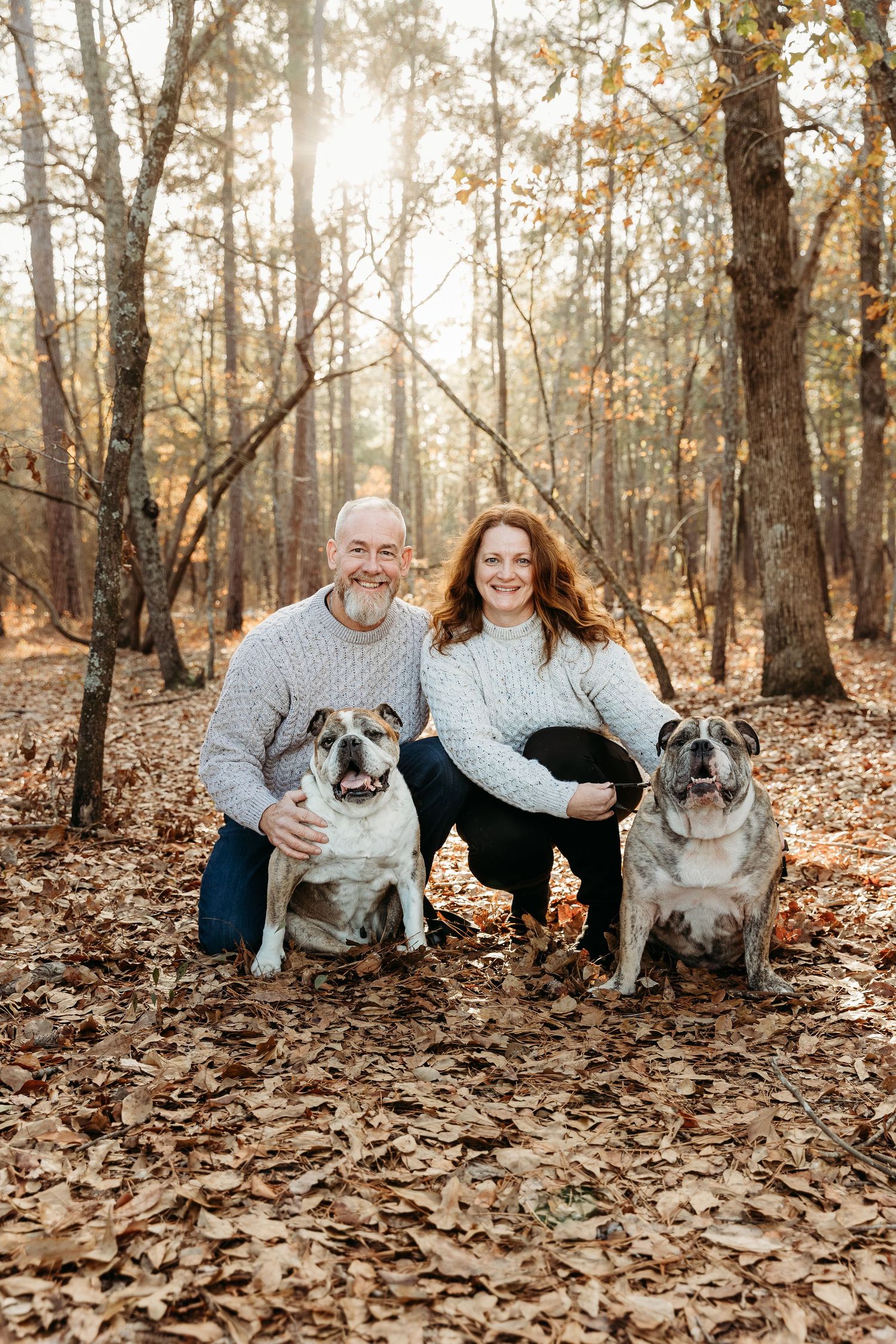 A couple sits with their dogs on autumn leaves in a sunlit forest setting.