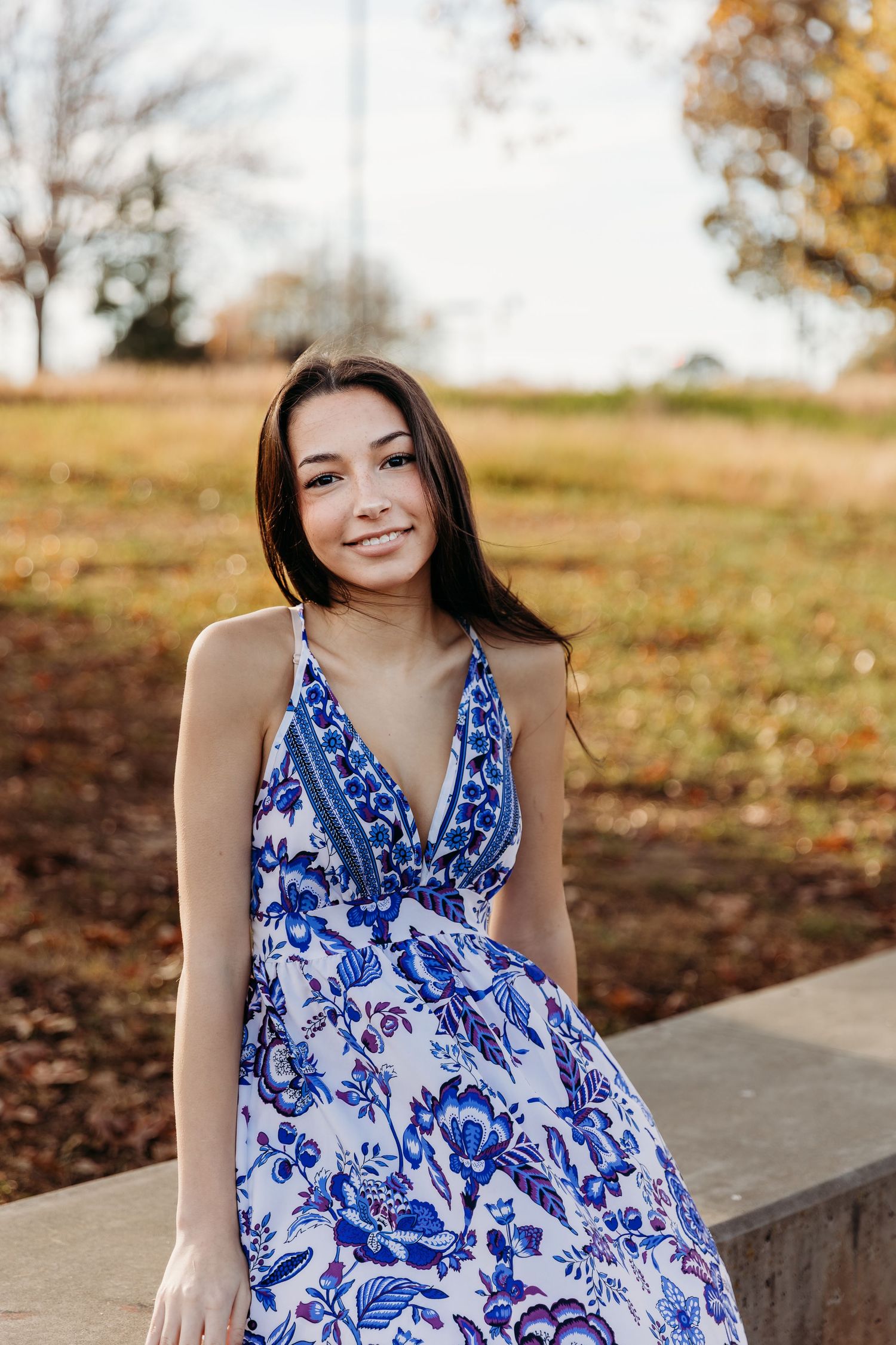 A person in a flowing blue and white floral dress poses outdoors during autumn.
