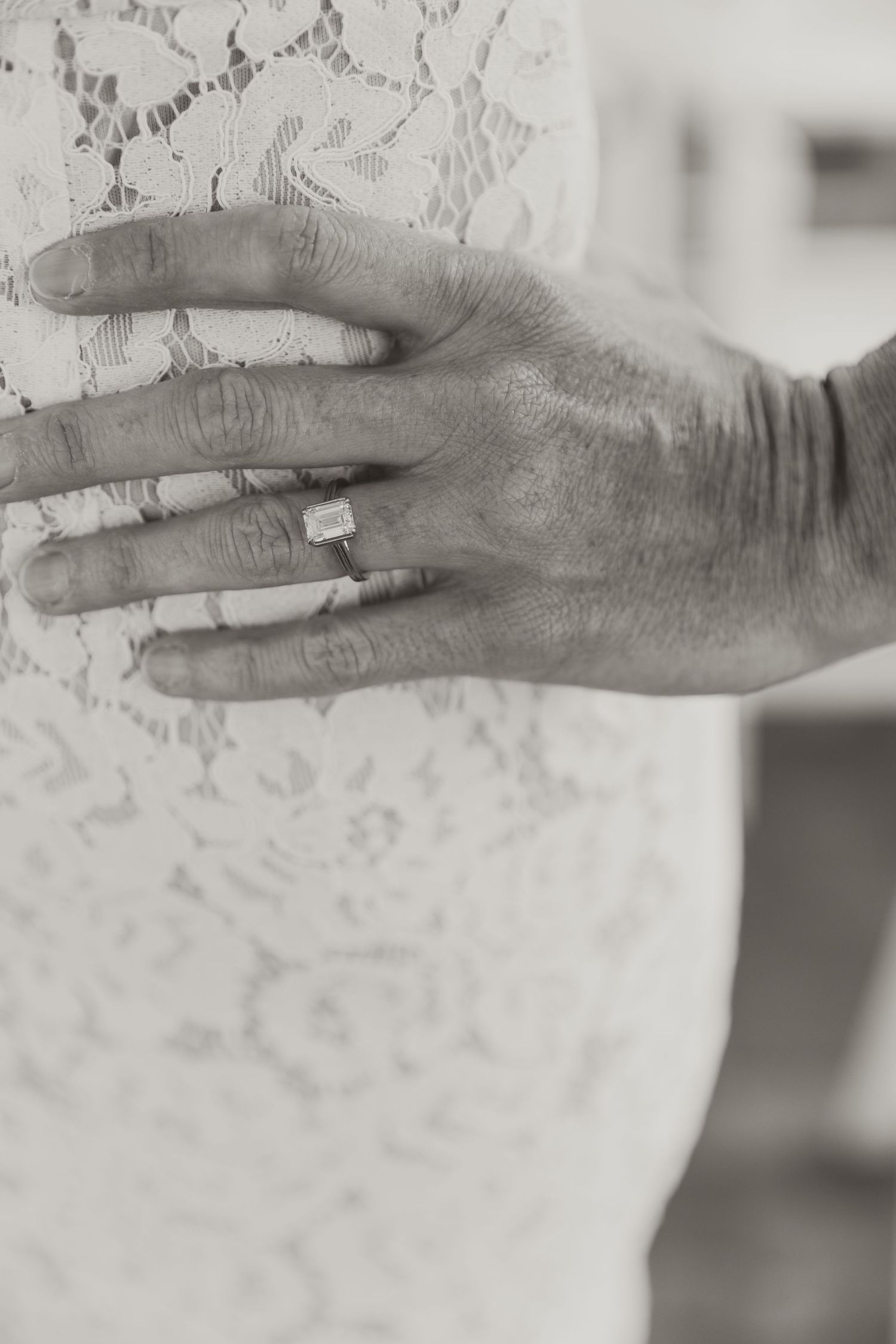 A close-up black and white photo of a hand wearing a diamond engagement ring against lace fabric.