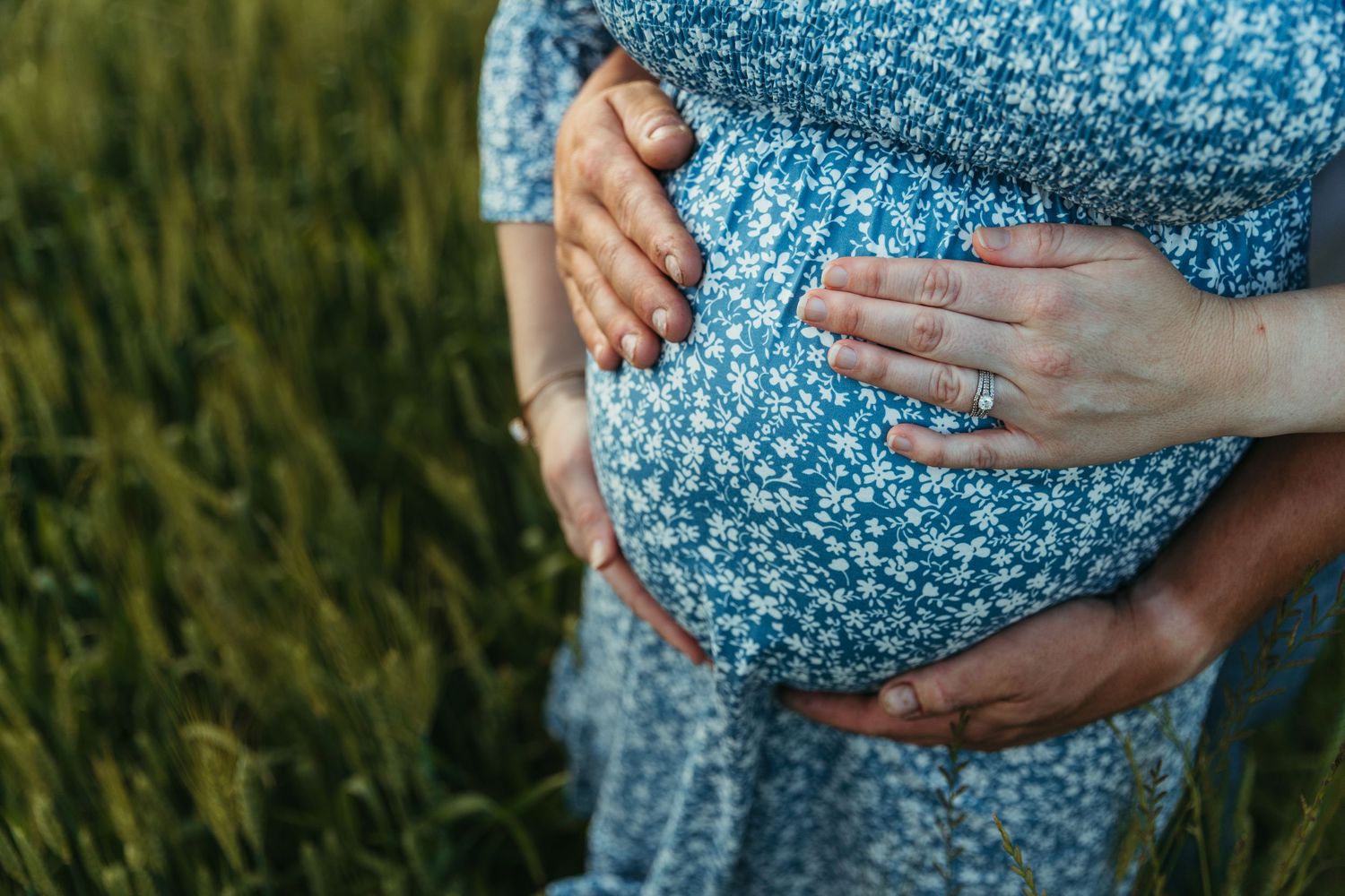 A pregnant woman in a blue floral dress cradles her baby bump while standing in a grassy field.