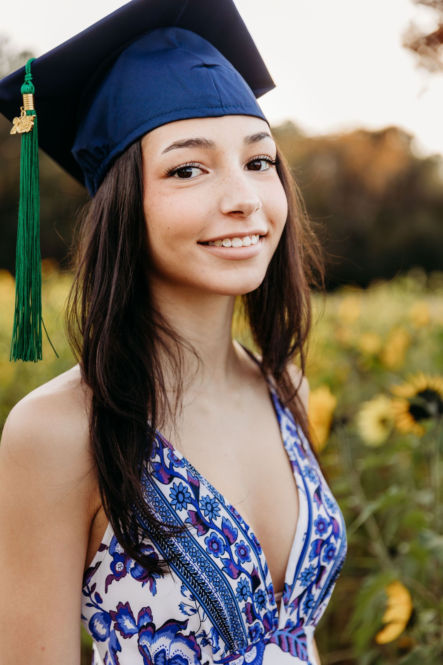 Graduate in blue and white floral dress poses in sunlit field wearing navy cap with green tassel.