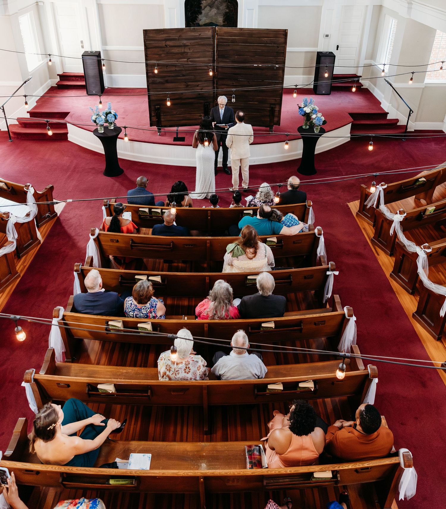 People sit in curved wooden pews with red carpeting inside a traditional church sanctuary.