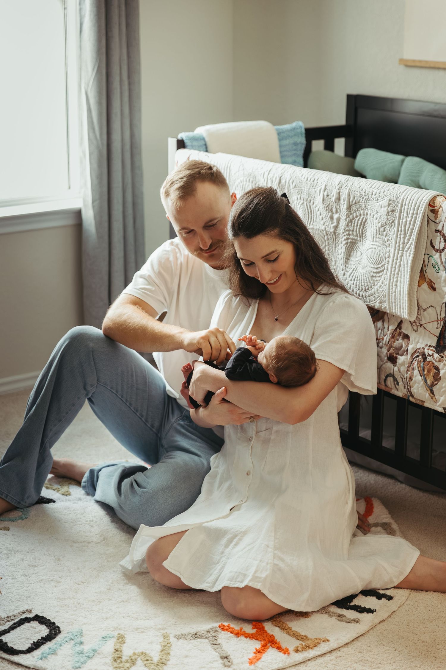 A couple in white clothing shares tender moments with their baby while sitting on the floor of a nursery.