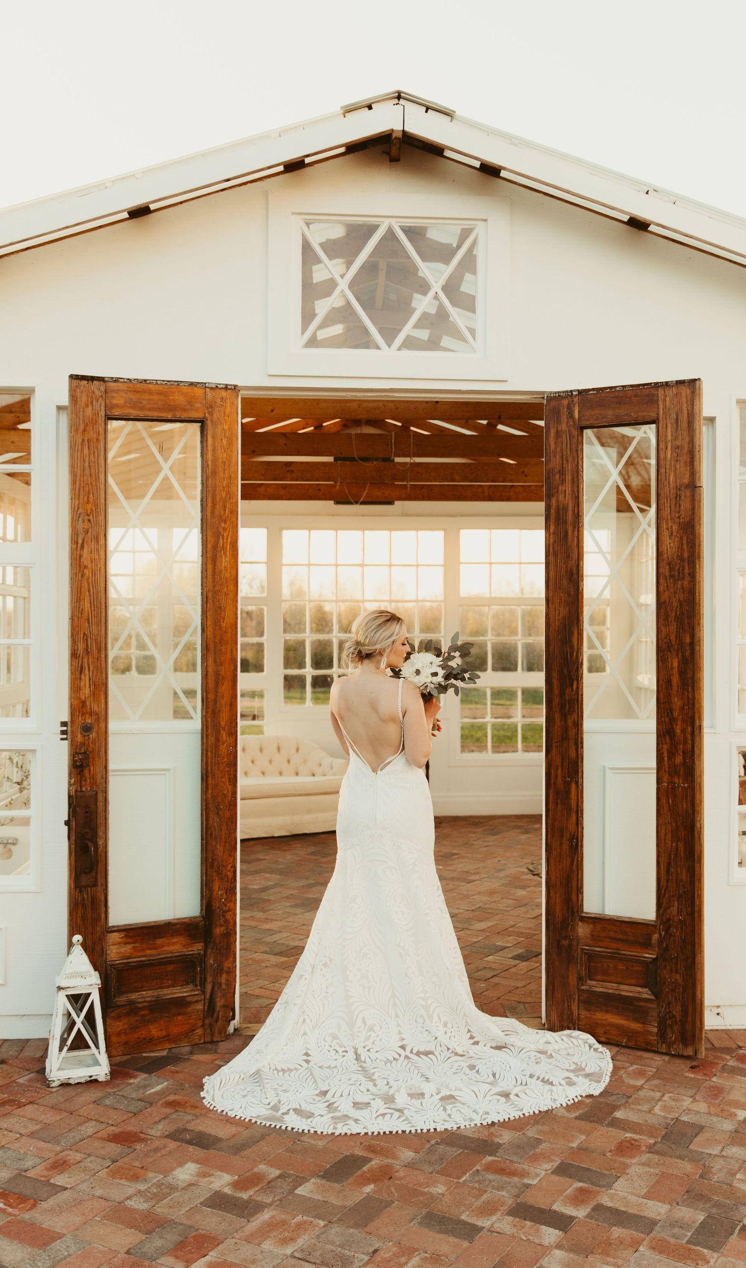 A person in a white dress stands in a rustic wooden doorway with large windows and natural light streaming in.
