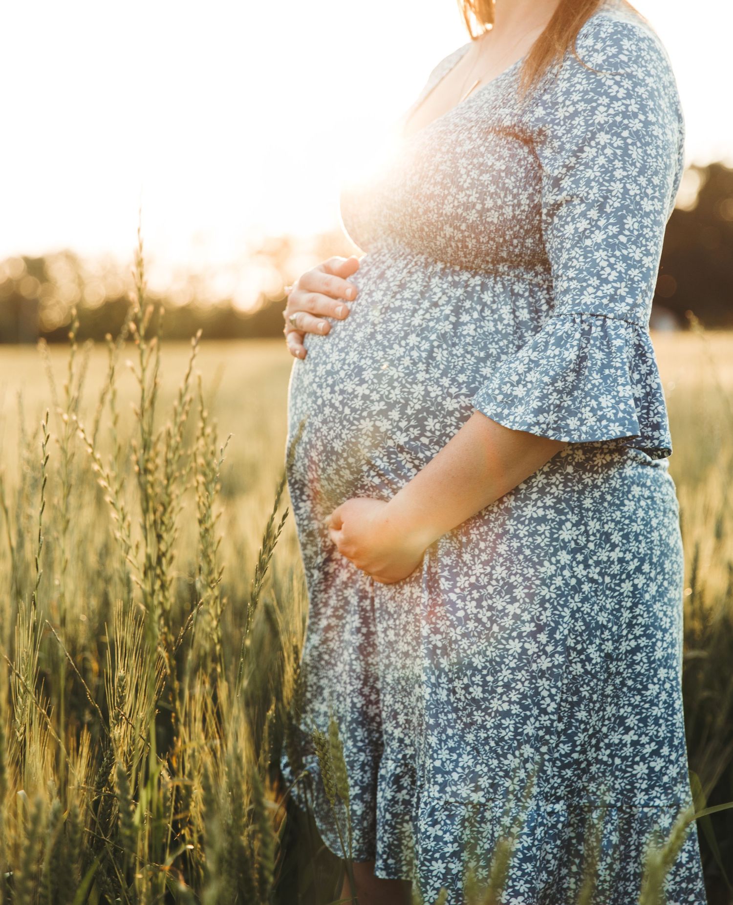 A pregnant woman in a floral dress stands in a golden wheat field at sunset, cradling her baby bump.