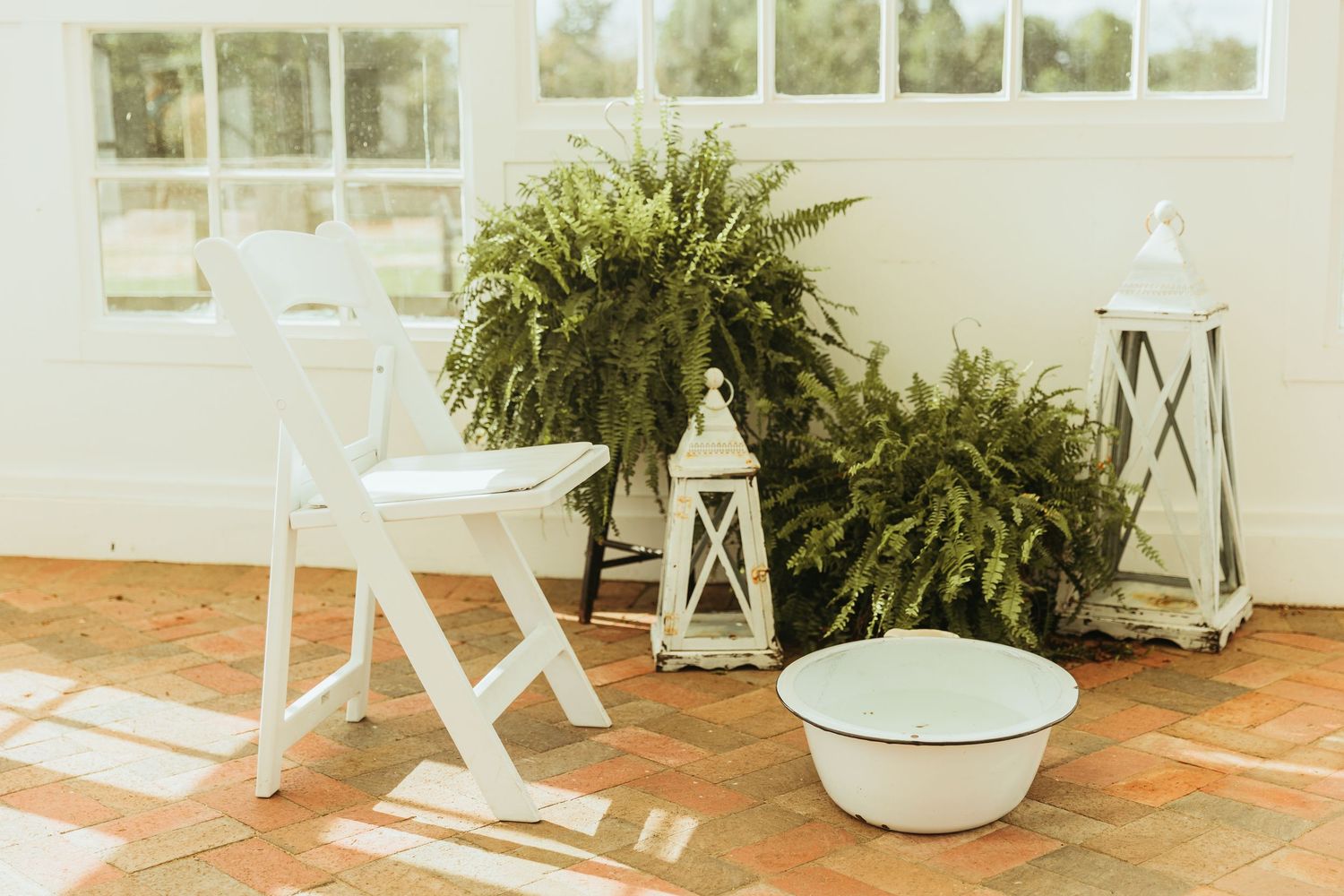 White wicker furniture and lush potted ferns create a cozy outdoor patio seating area in natural sunlight.