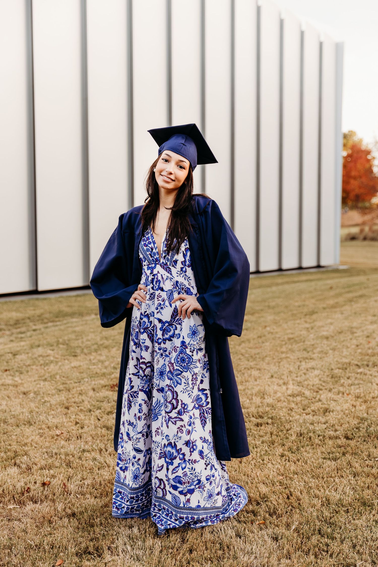 Graduate in blue and white floral dress poses with graduation cap and gown in outdoor setting with autumn trees.