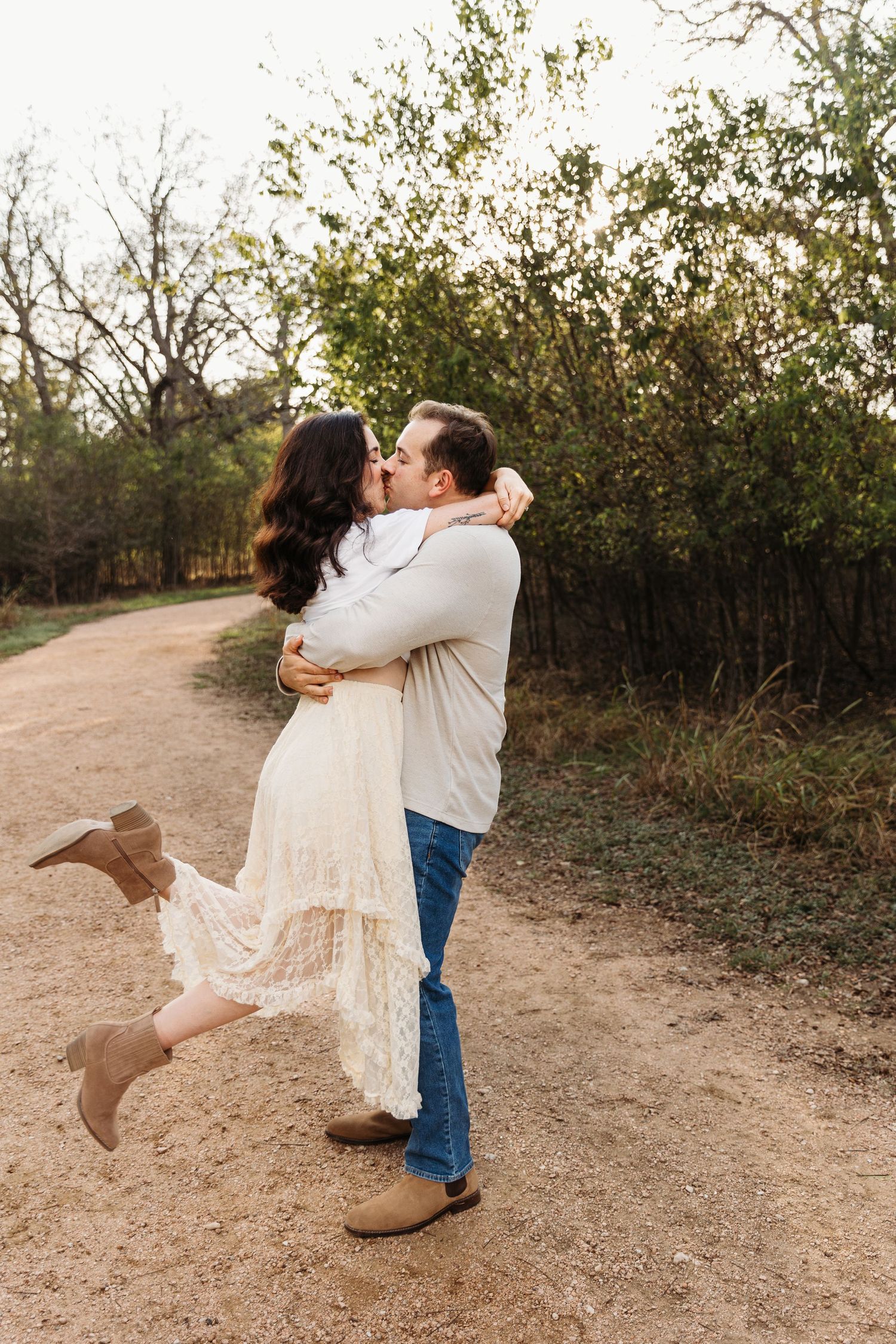A romantic couple in casual attire dances and spins together on a dirt path surrounded by autumn foliage.