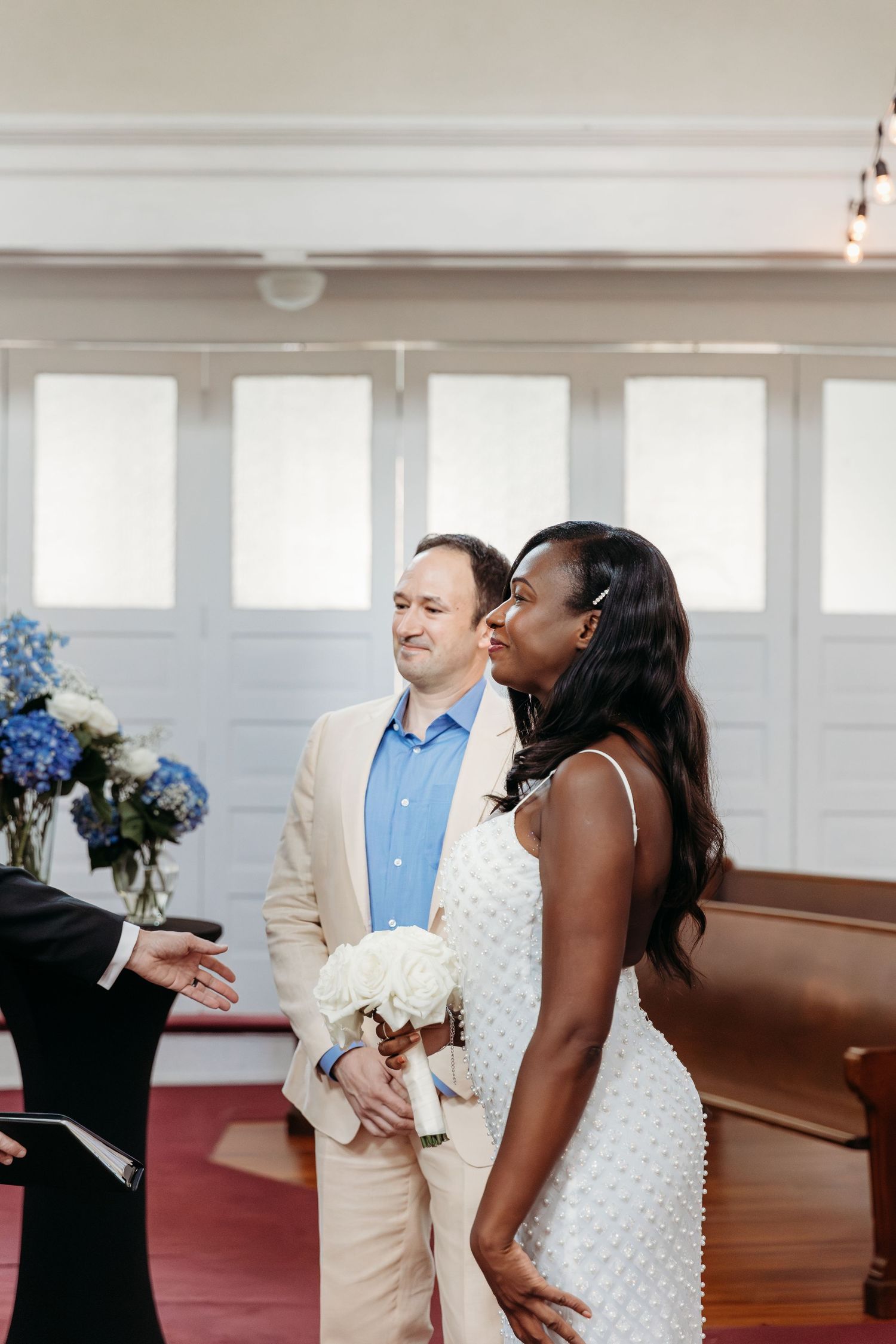 A couple in formal attire stands together in a church with white walls and red carpeting during a ceremony.