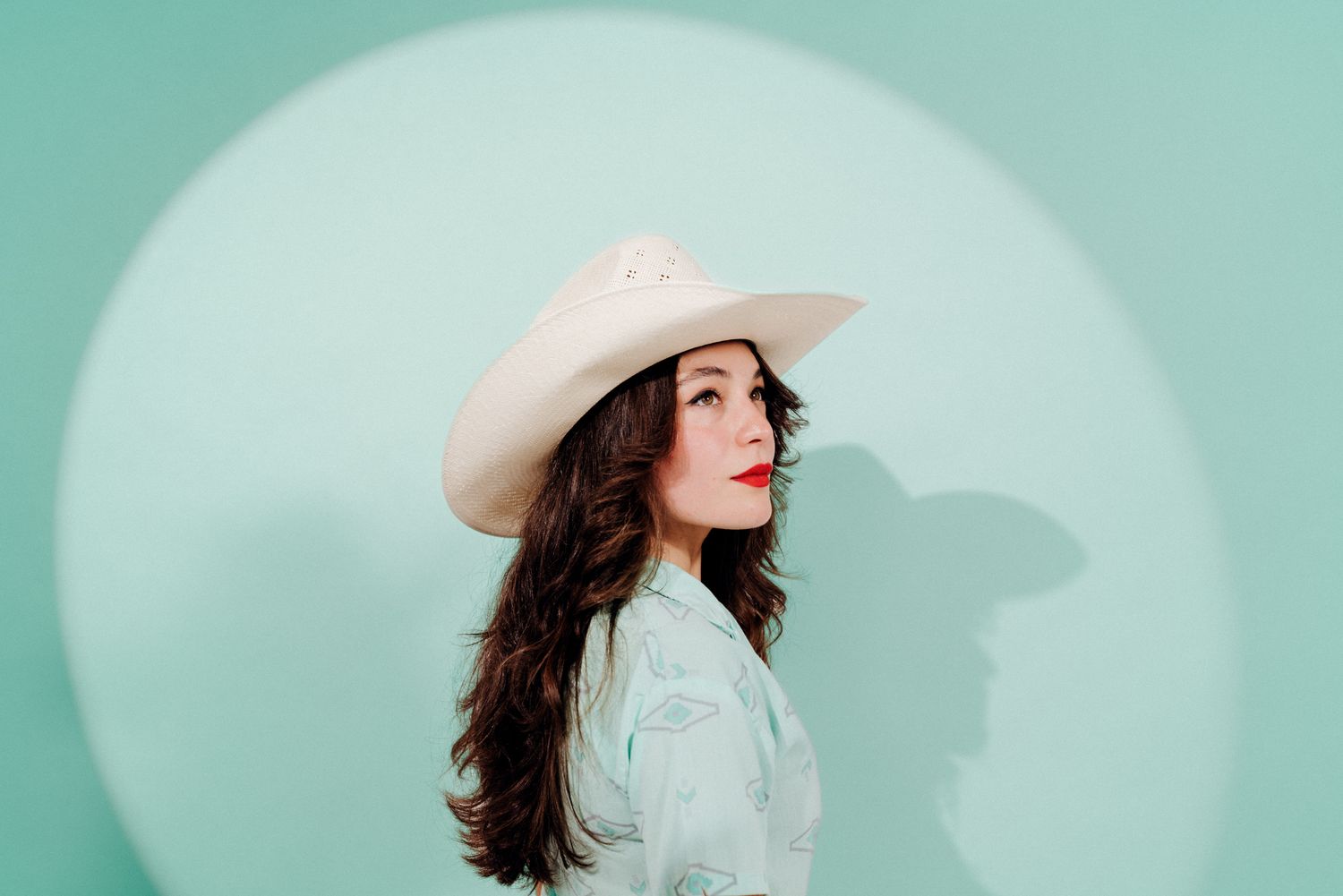 A portrait of a model in a white cowboy hat against a mint green background with dramatic side lighting.