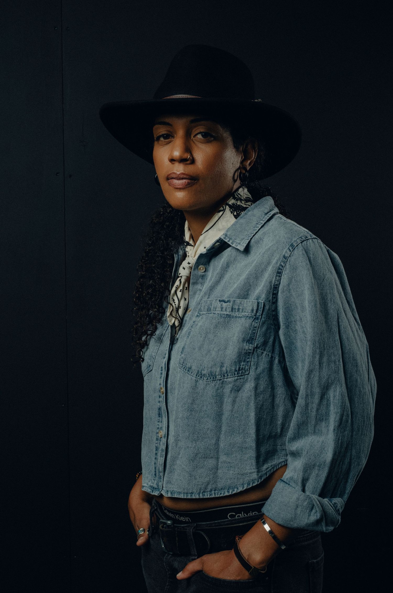 Portrait against dark background showing person in black hat, denim shirt and dark pants in western-inspired style.