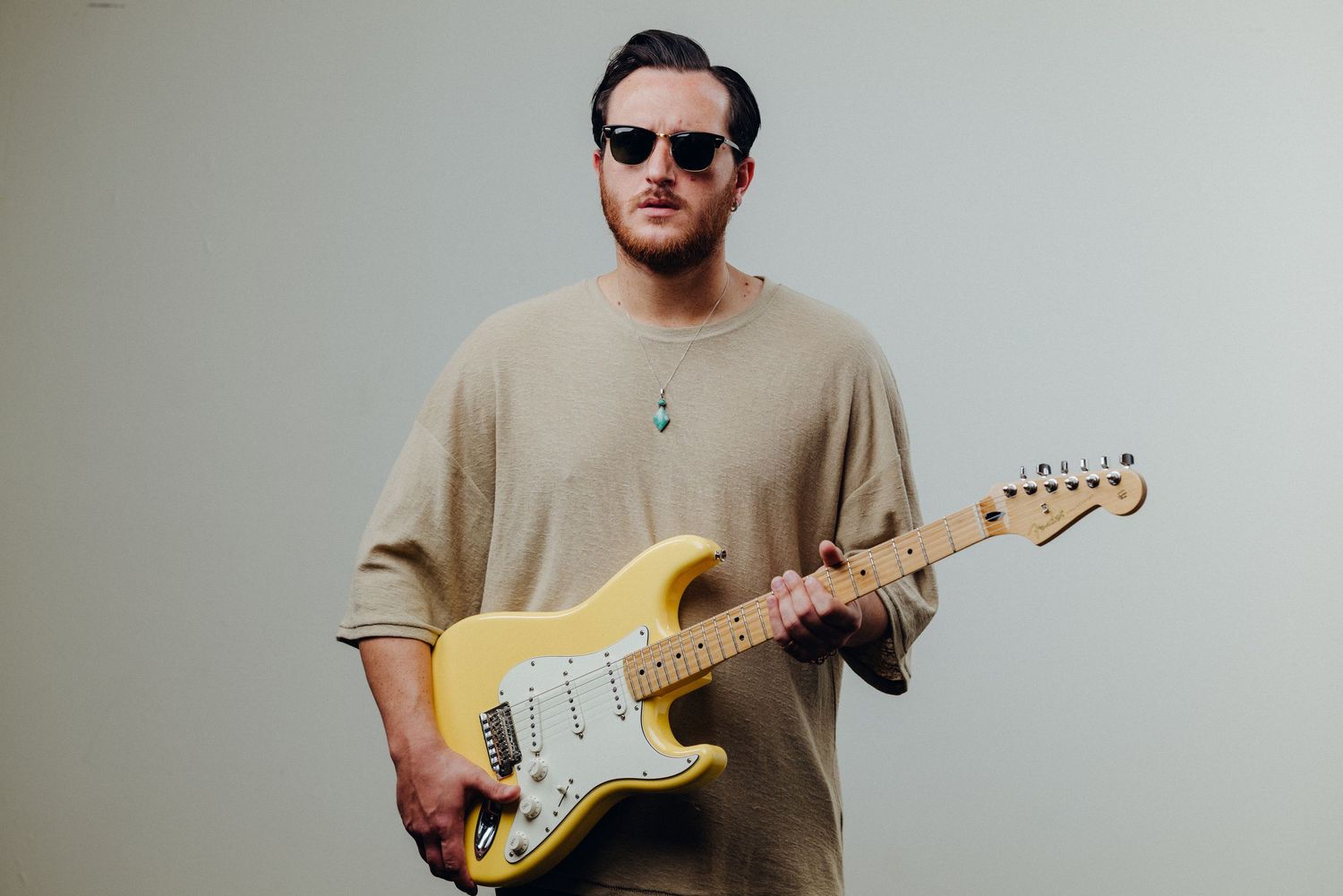 Musician in beige shirt playing electric guitar against neutral backdrop.