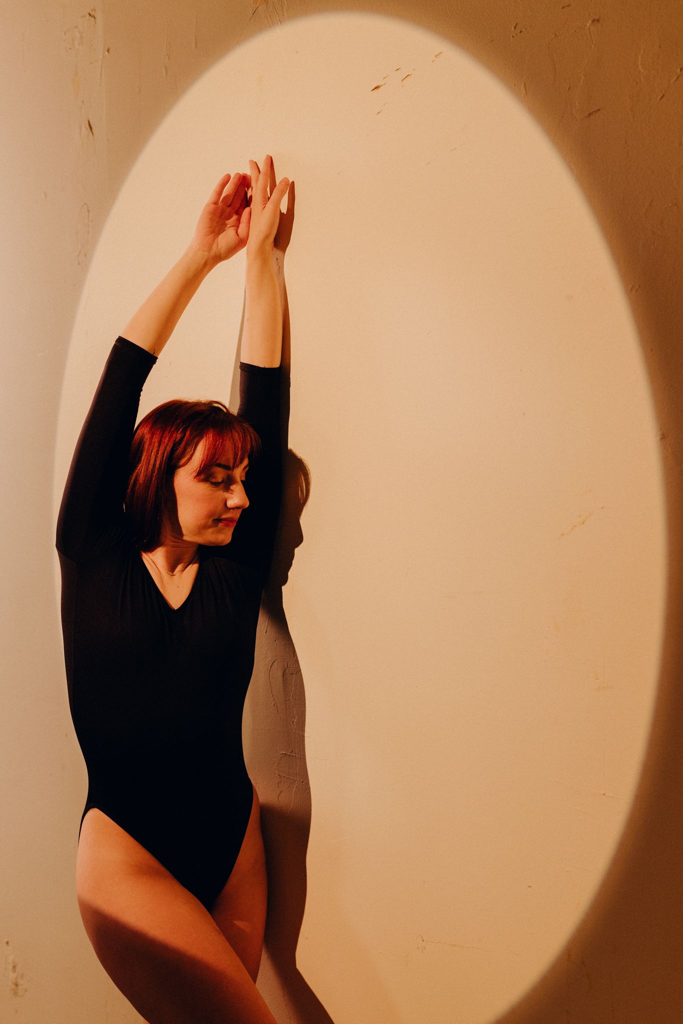 Silhouetted figure performs expressive dance movements against beige backdrop with circular spotlight.