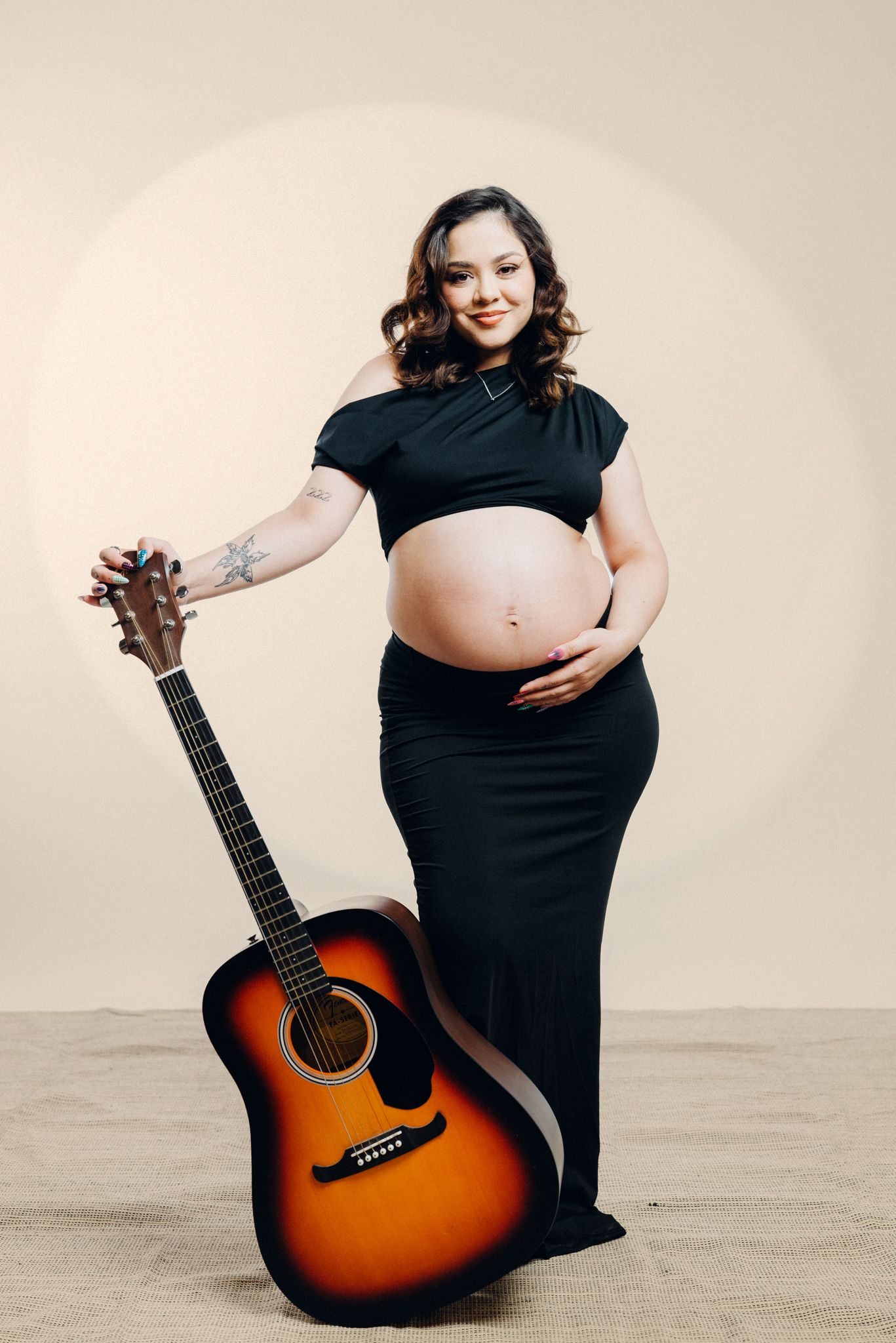Maternity photoshoot featuring acoustic guitar and black dress against neutral background.