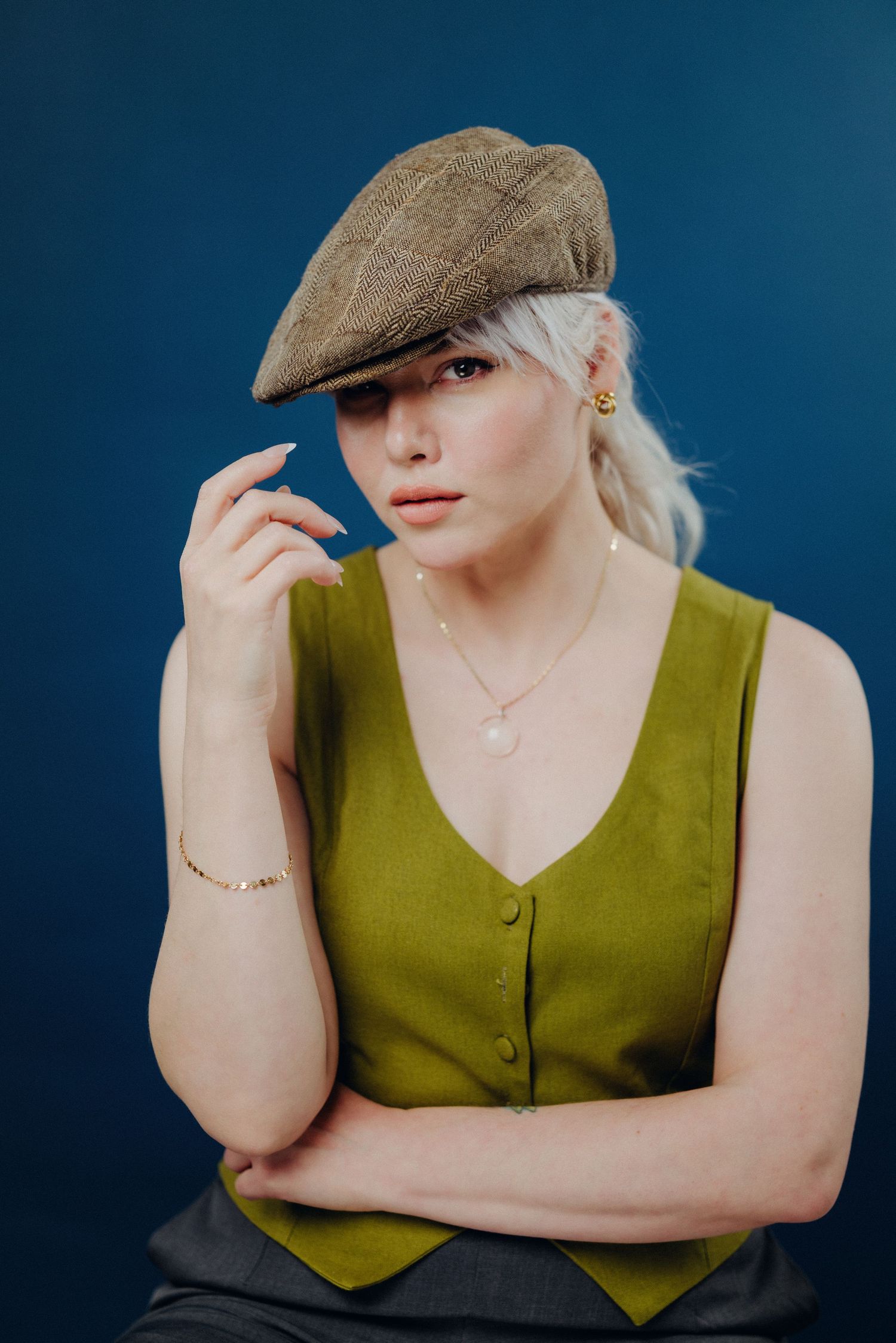 Stylish figure in olive vest and newsboy cap poses dramatically against deep blue backdrop.