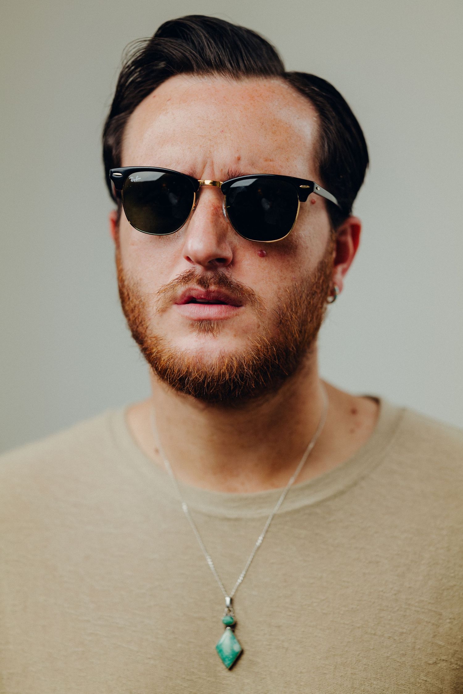 Close-up of person wearing sunglasses and turquoise pendant necklace against light background.