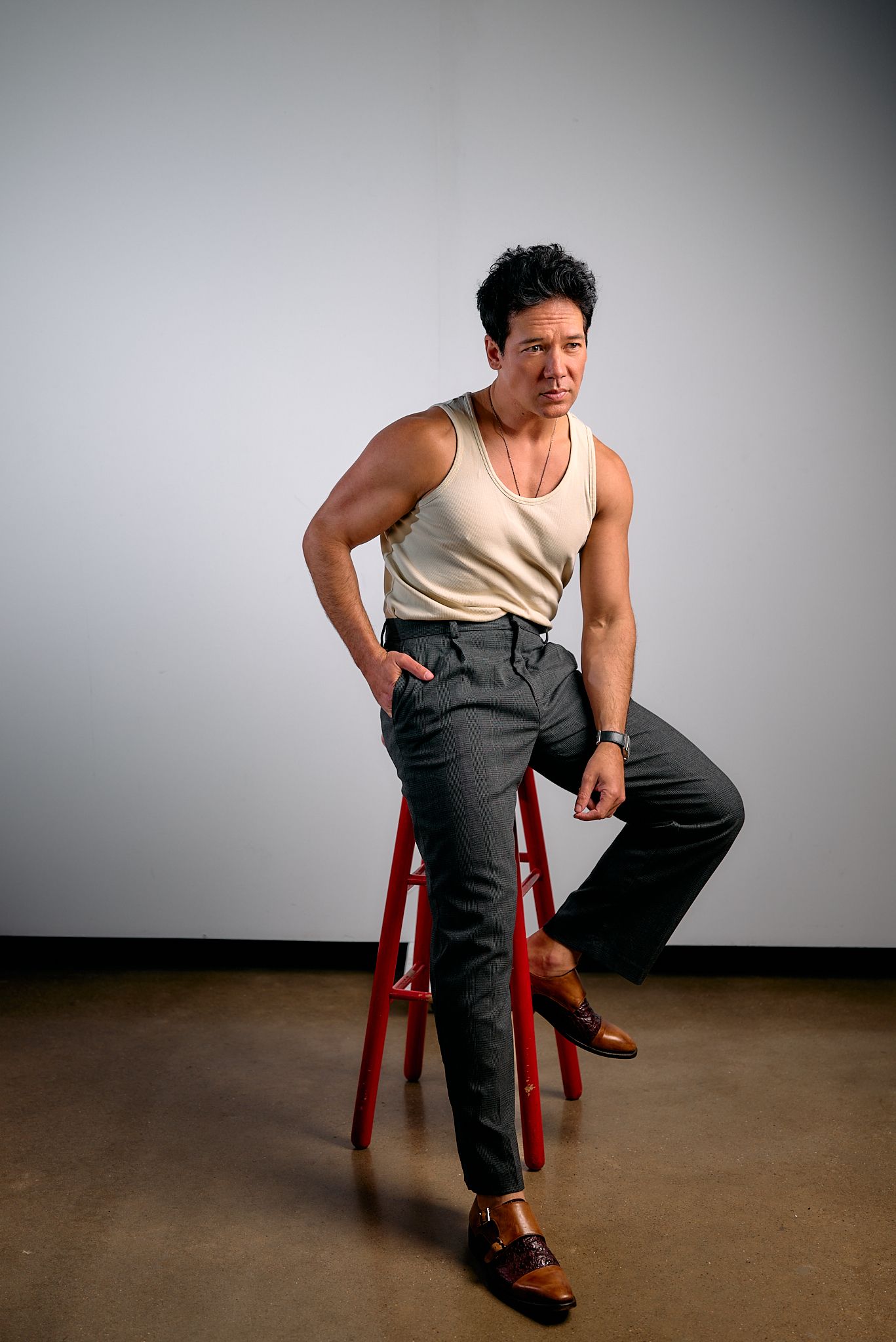 A person sits on a red stool wearing a white tank top and dark pants in a sequence of four similar poses.