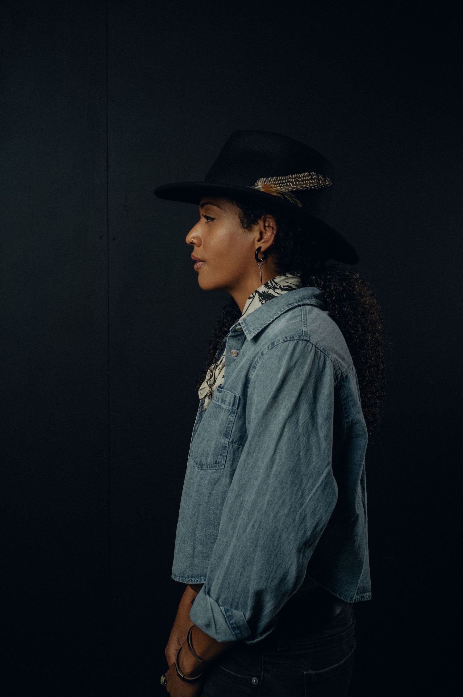A person in a denim jacket and baseball cap poses against a dark background in a fashion photography series.