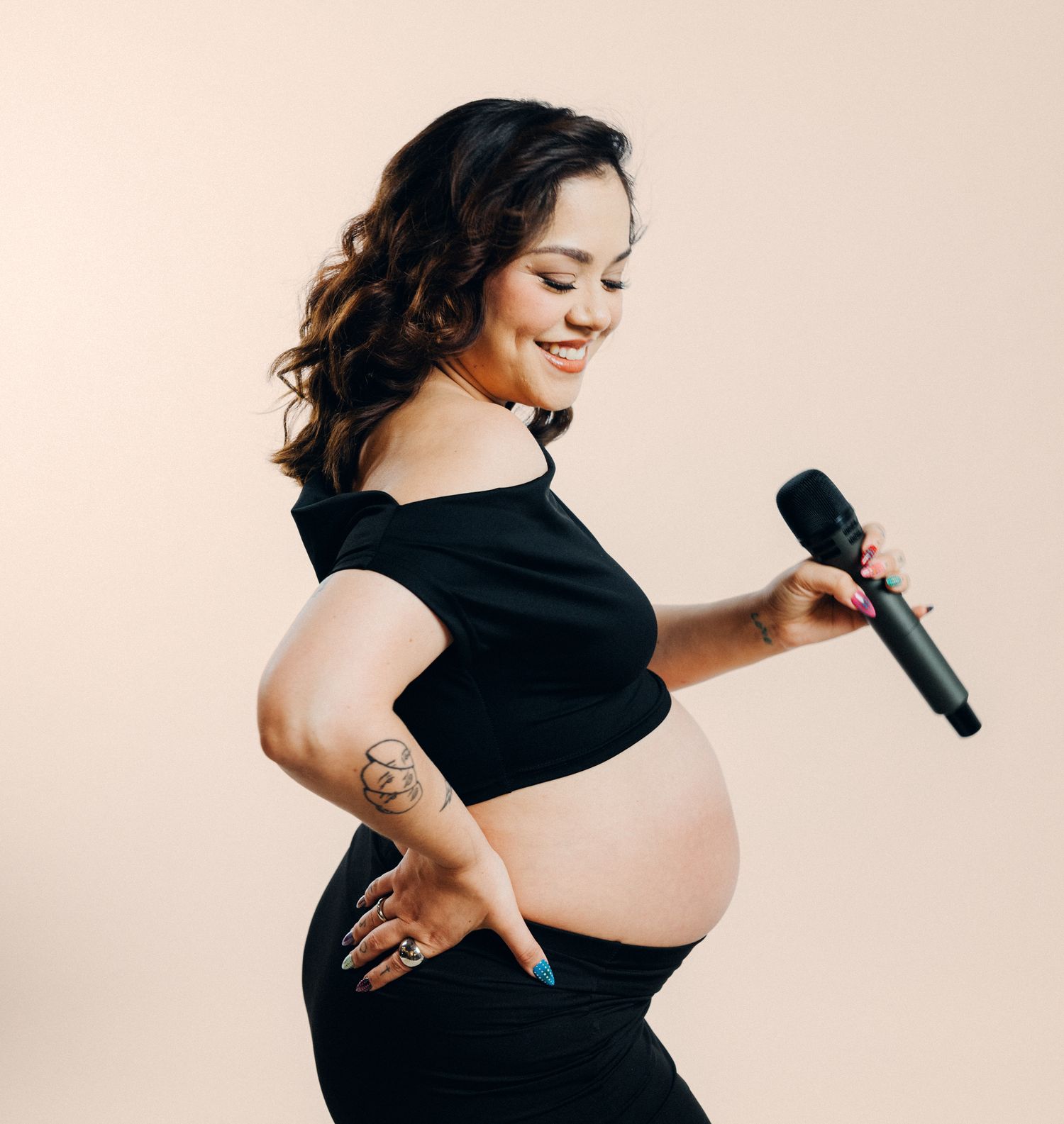 Woman in black dress holds microphone during maternity photoshoot against beige backdrop.