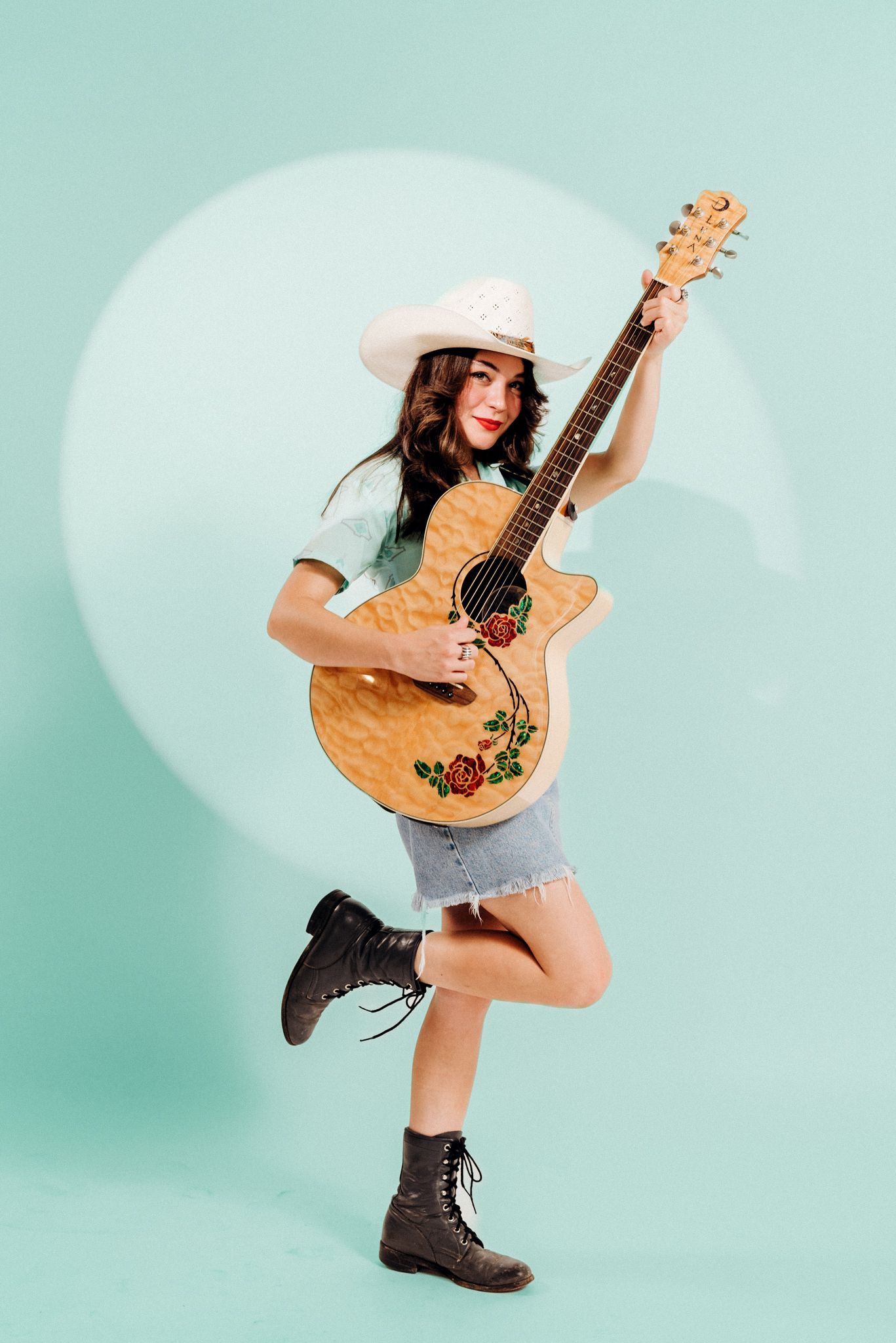 Energetic musician poses with acoustic guitar against mint green background wearing cowboy boots.