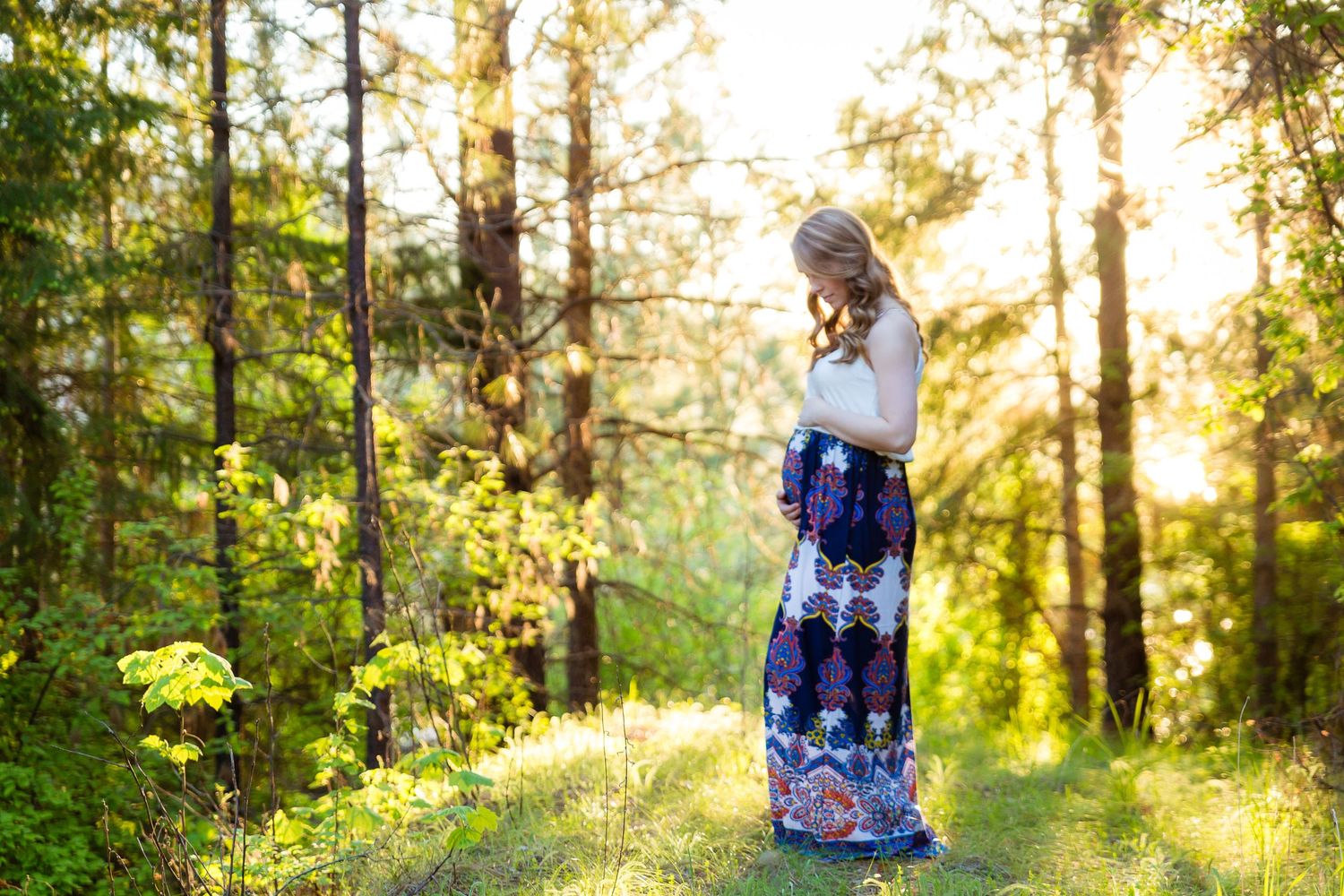 Woman in flowing maxi dress stands in sunlit forest clearing.