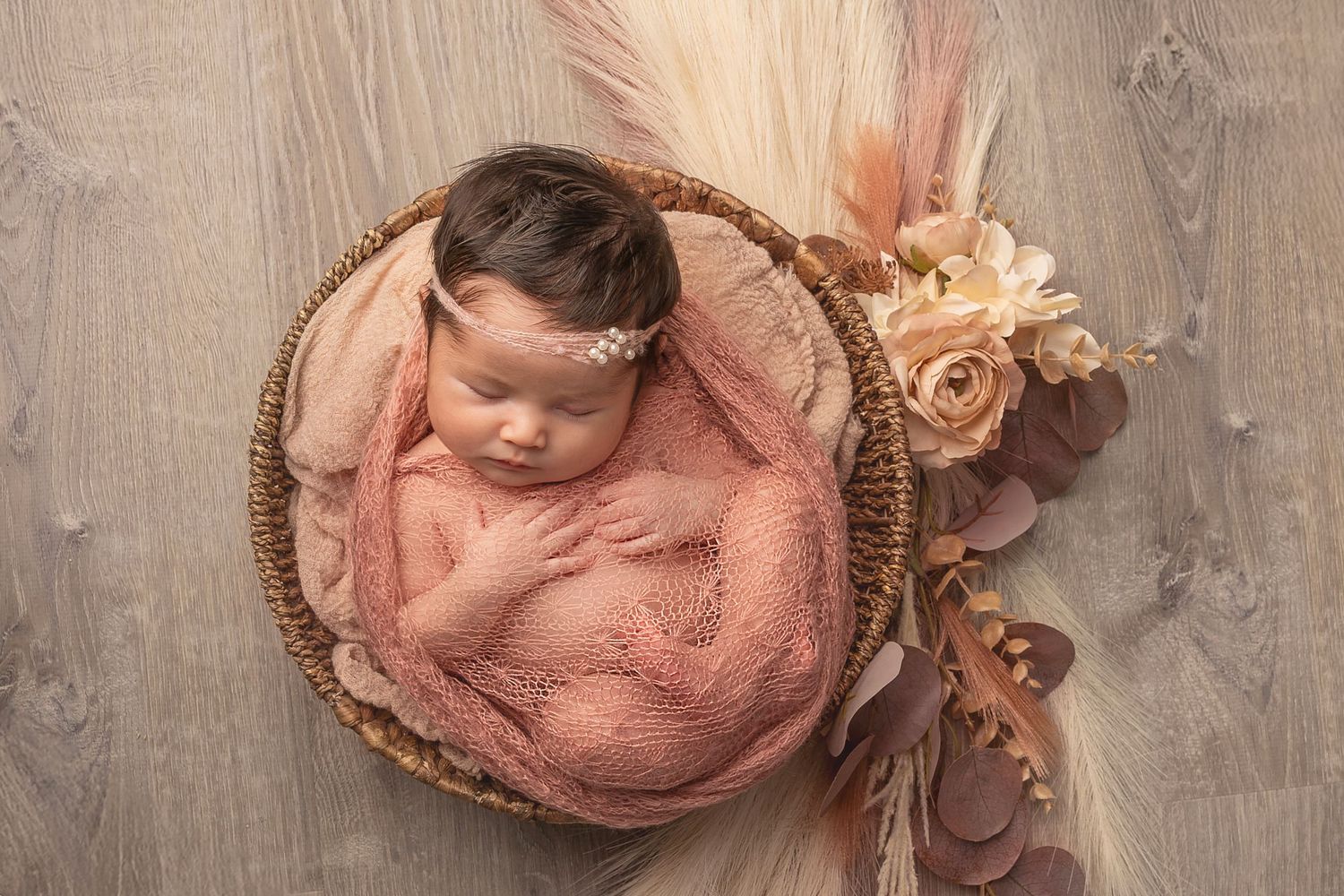 Sleeping baby wrapped in soft pink fabric lying in round wicker basket with peach-colored floral decorations.