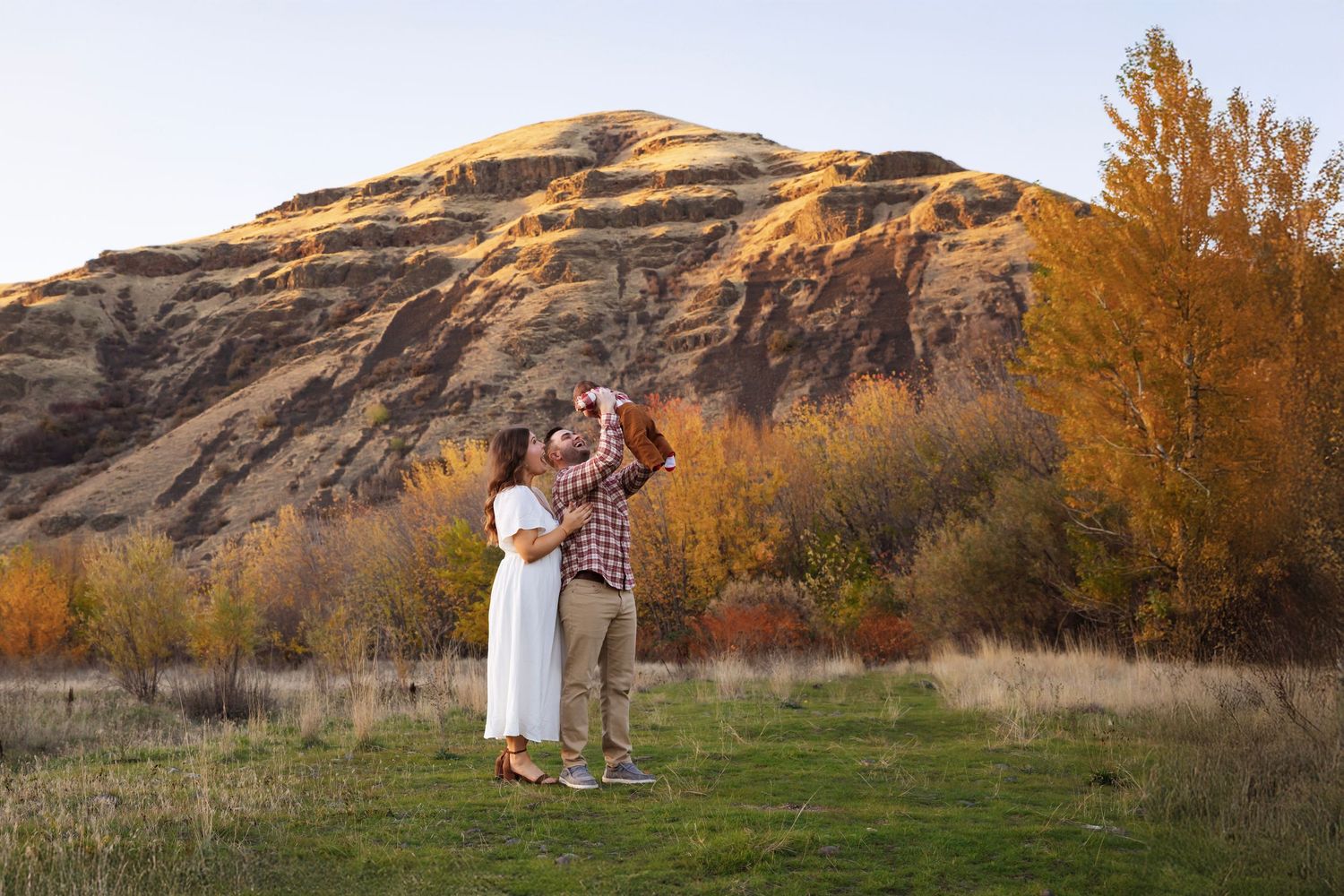 Couple embraces in golden autumn field with mountains and trees creating romantic fall engagement photo backdrop.