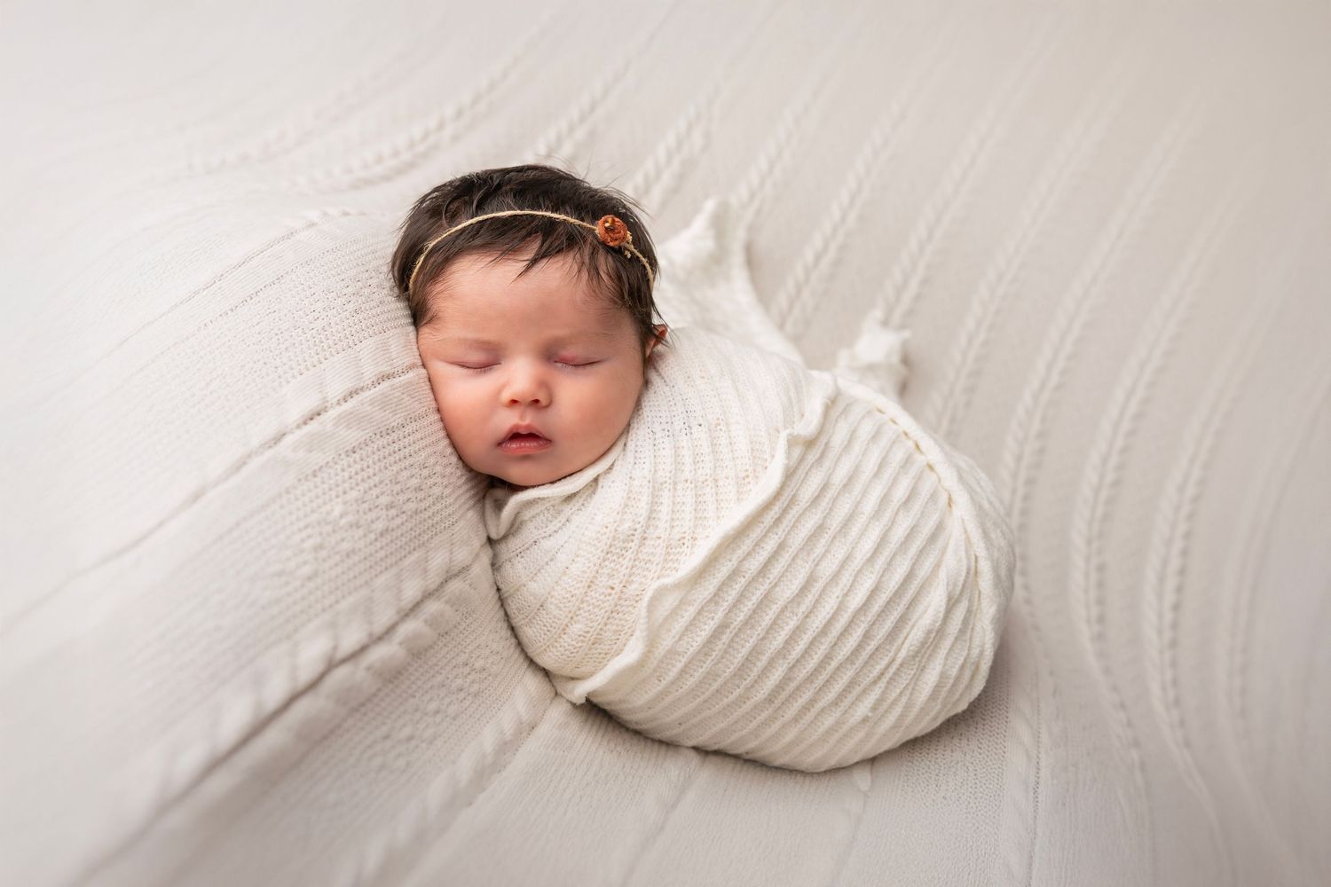 Sleeping newborn baby wrapped in soft white swaddle blanket on white textured surface.