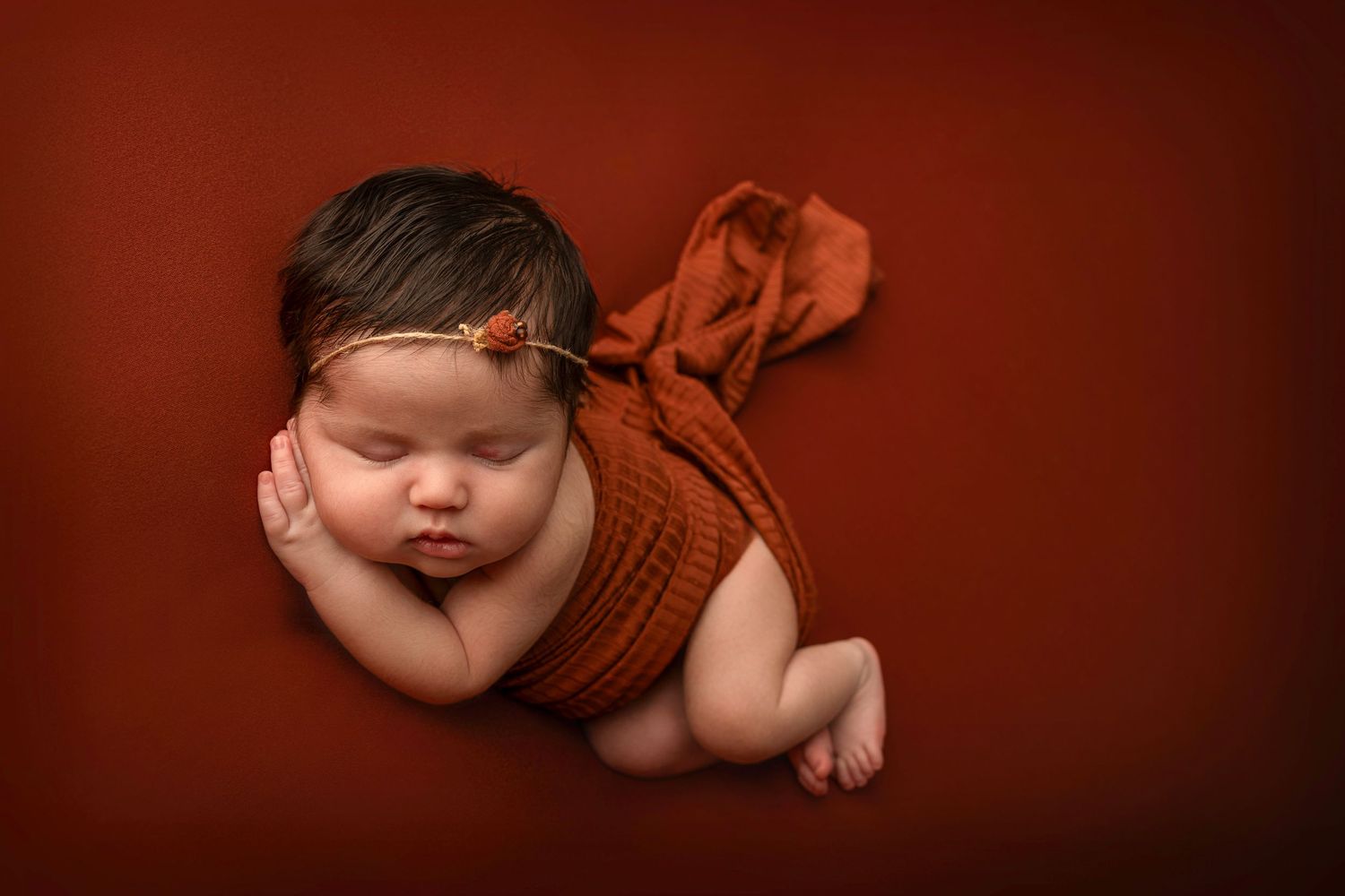 Adorable sleeping newborn baby wearing brown knit outfit against dark red background in artistic portrait.