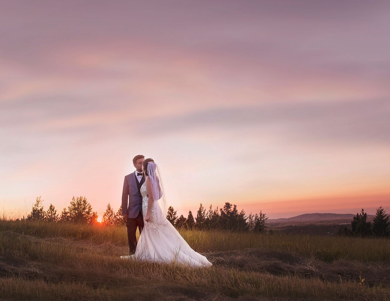 Newlywed couple stands together on hilltop during dramatic sunset.