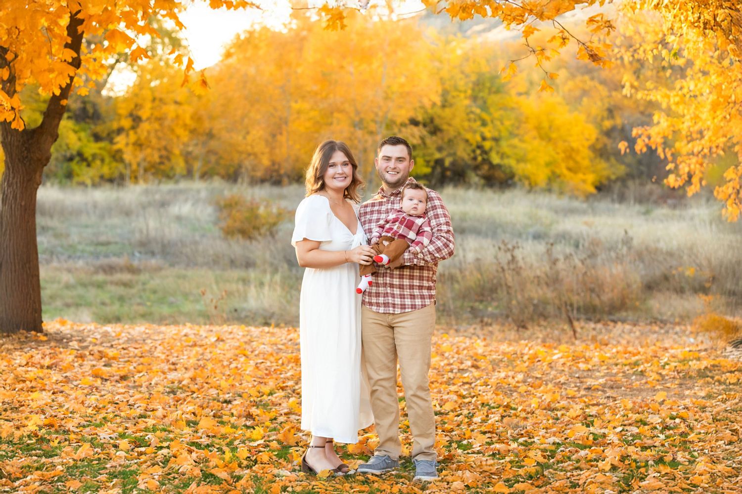 A couple stands together in a golden autumn setting with fallen leaves and warm sunlight filtering through trees.