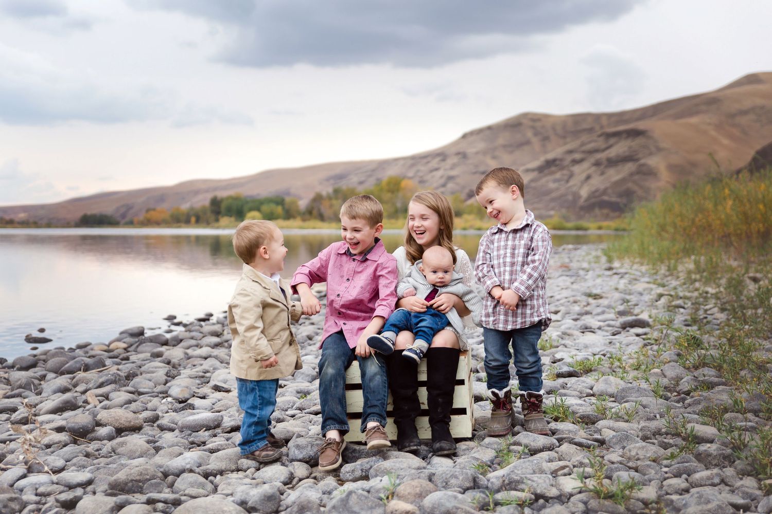 Children sit together on rocky shoreline with mountains in background.