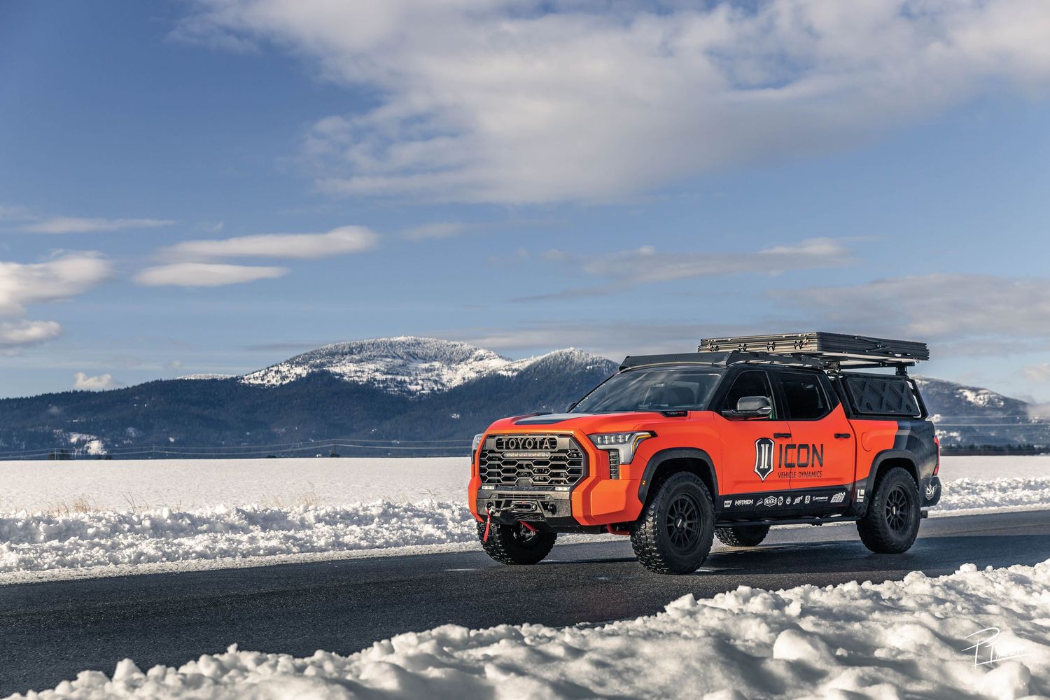 A red off-road vehicle drives across a snowy mountain landscape with dramatic cloudy skies in the background.