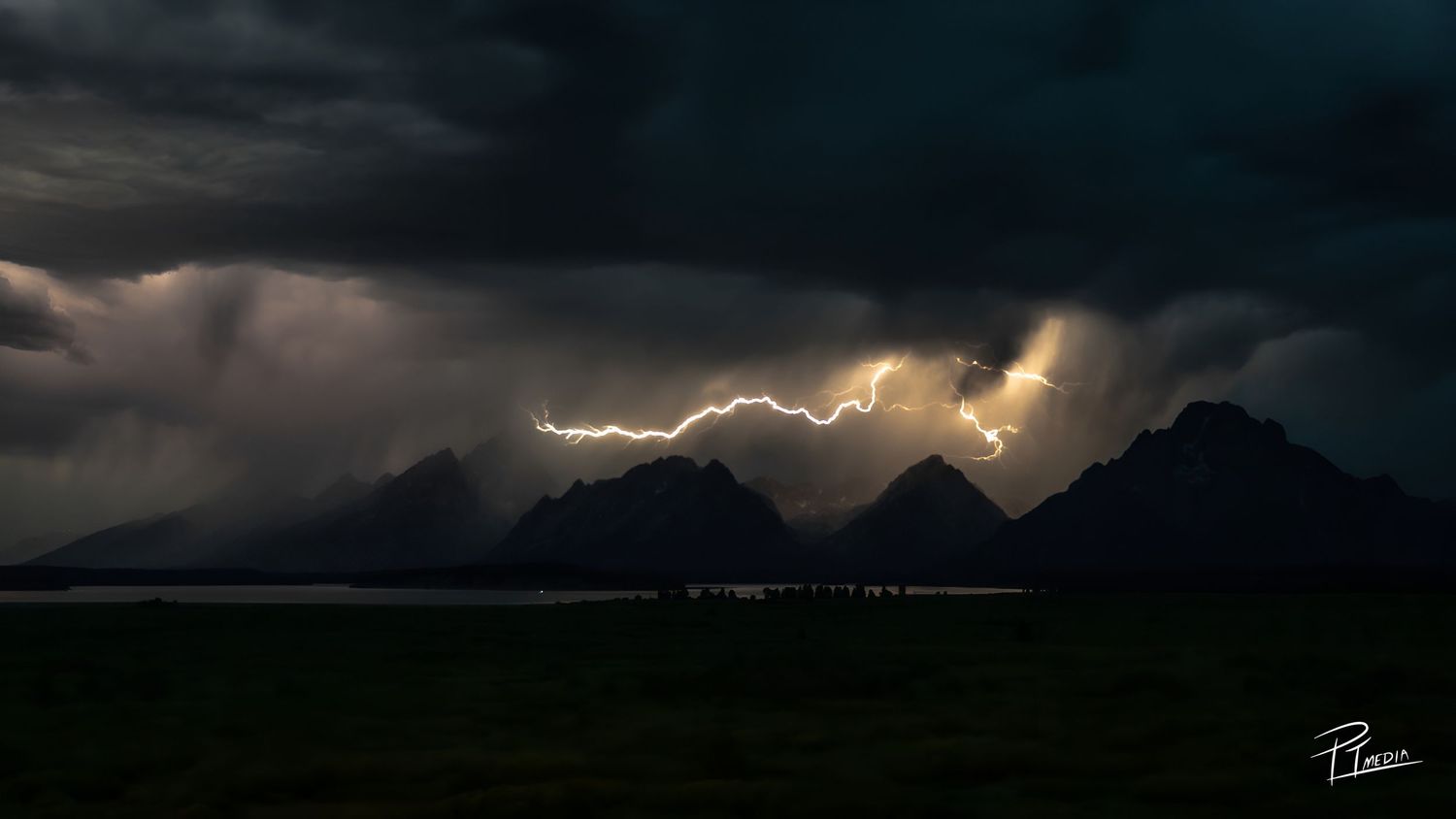 Lightning illuminates dark stormy skies over mountain ranges in a dramatic panoramic landscape photograph.