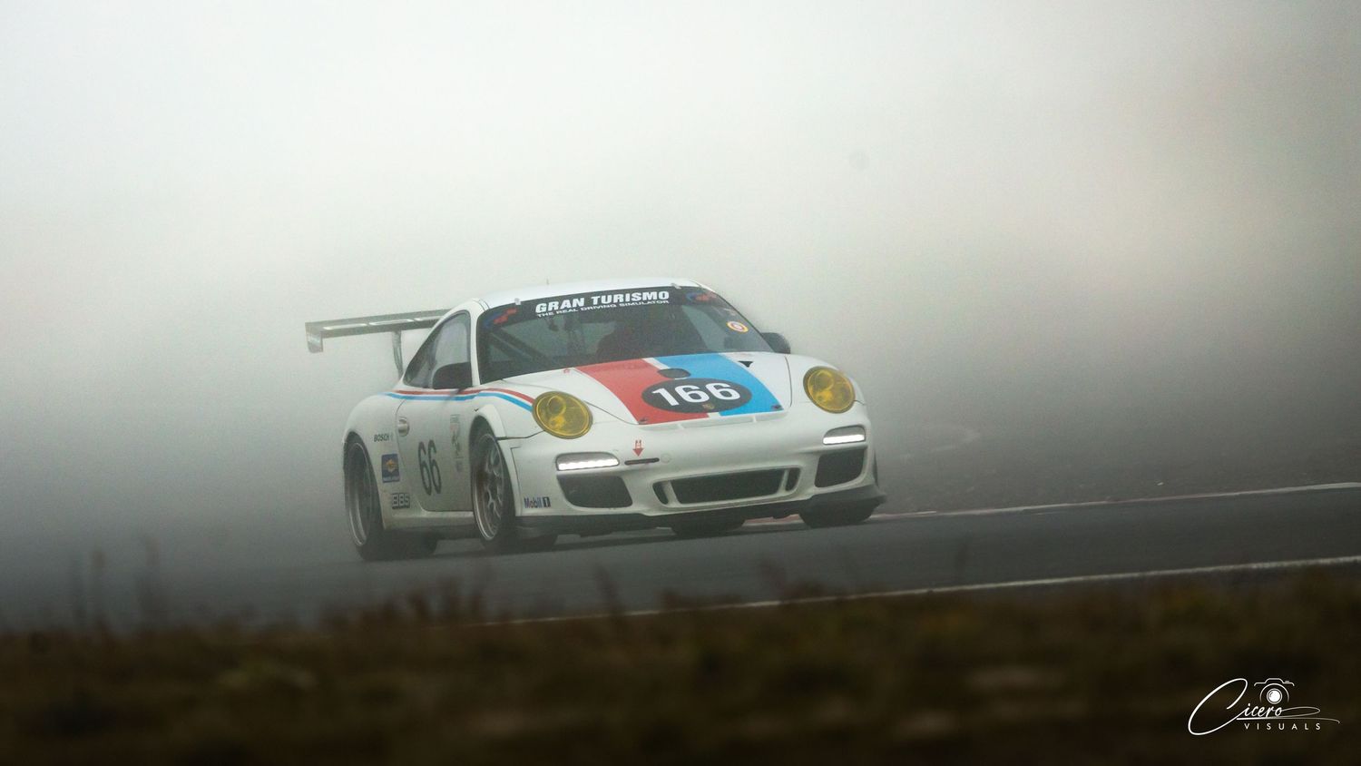 White Porsche 911 GT3 race car driving through foggy conditions on a race track.