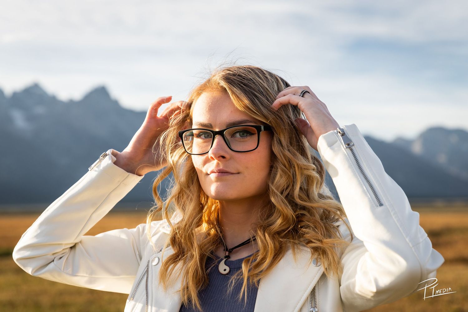 Person in glasses poses outdoors with mountains and dramatic sky in the background during golden hour.