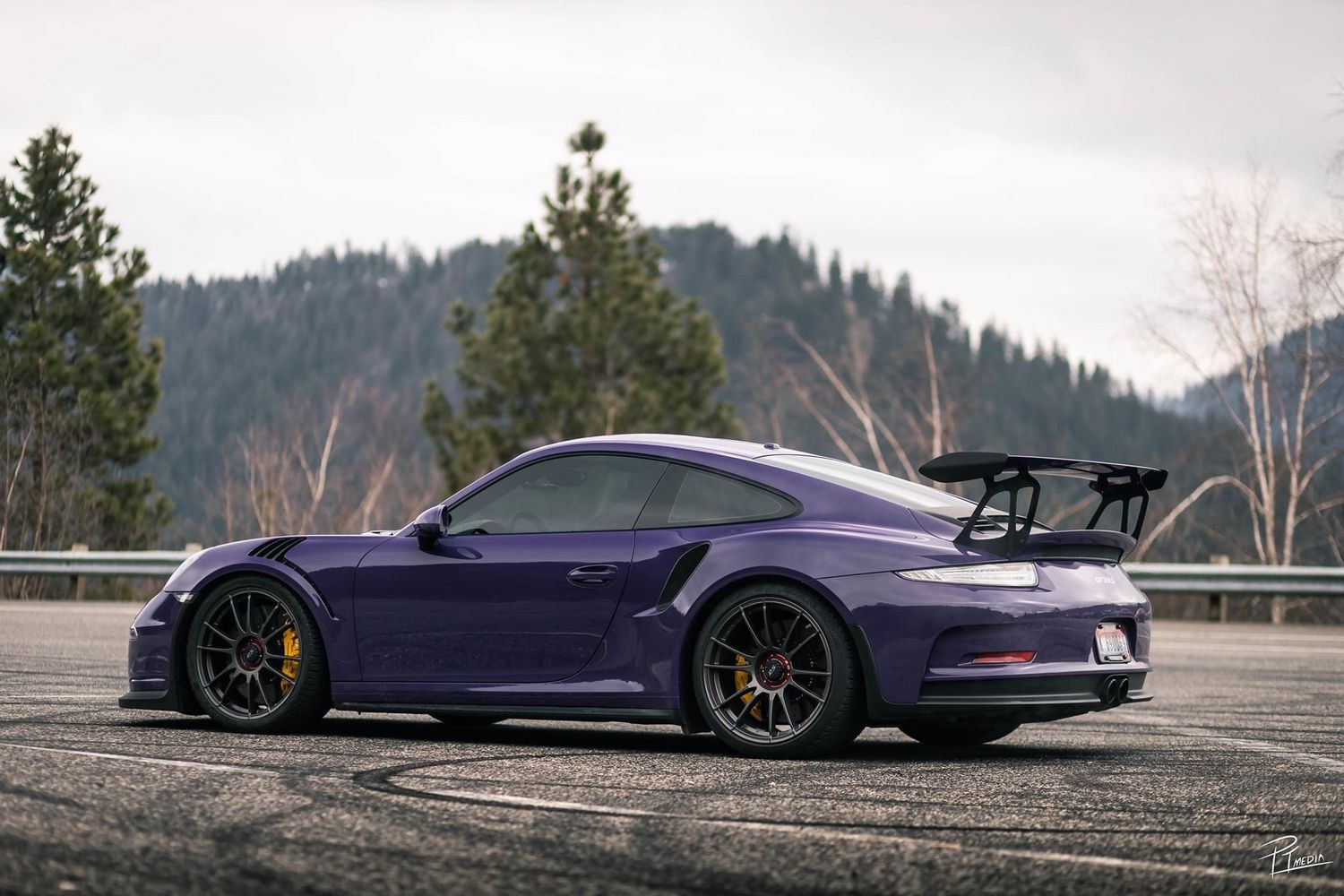 Purple Porsche GT3 RS with black wheels and large rear wing parked on mountain road with forest backdrop.