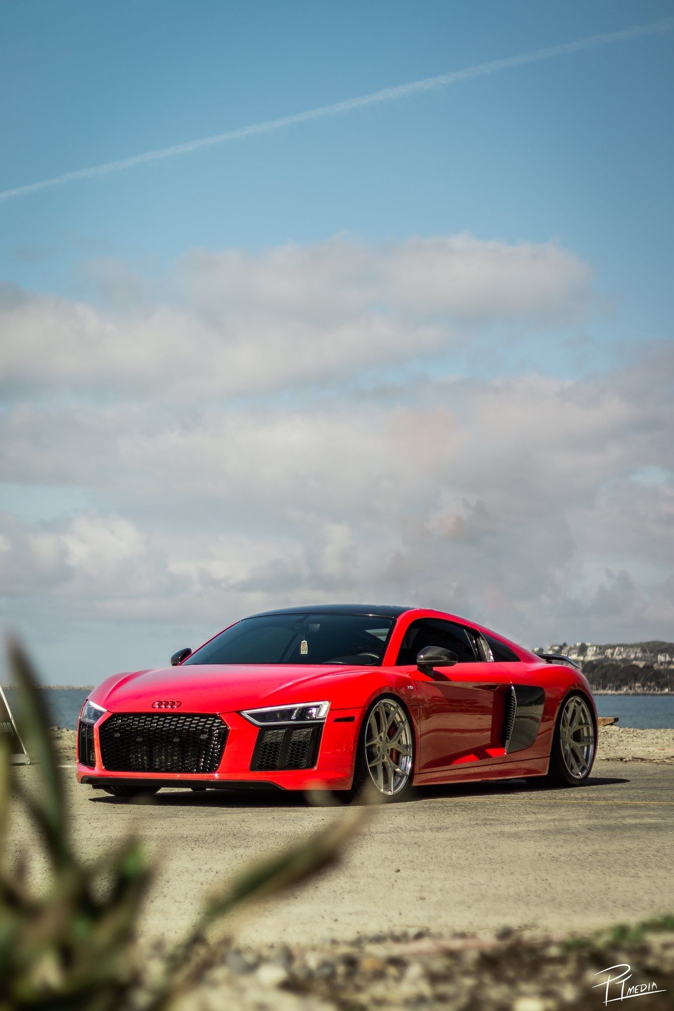 Red Audi R8 sports car parked on a sandy beach with ocean and cloudy sky in the background.