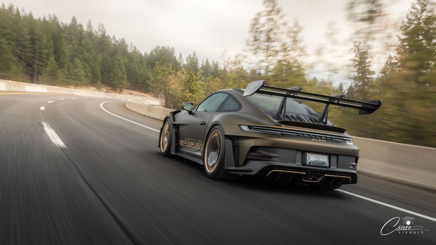 A black sports car with a large rear spoiler speeds along a winding forest road in dramatic motion blur.