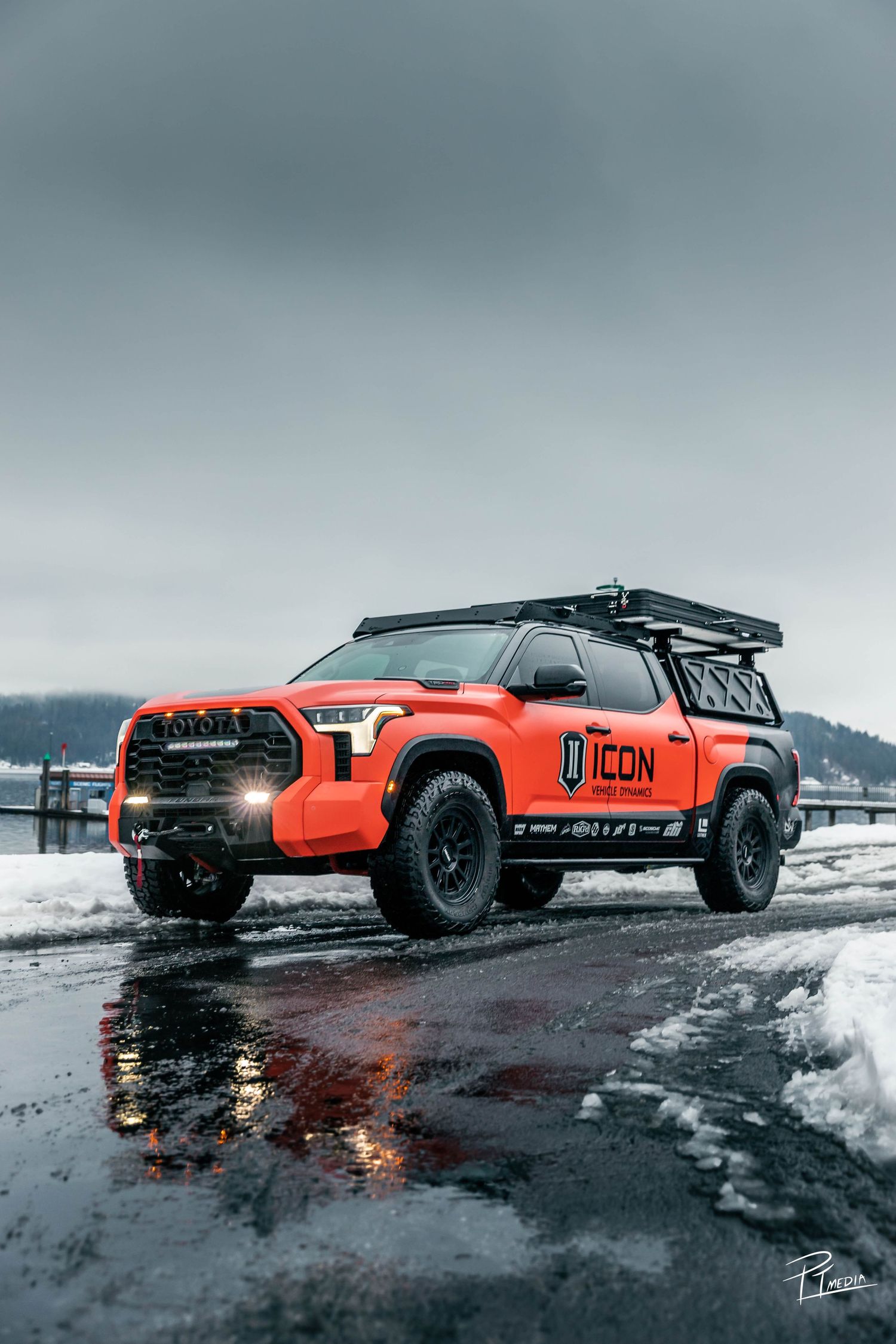 Orange sports car driving through snowy terrain with dramatic moody sky and reflective wet ground.