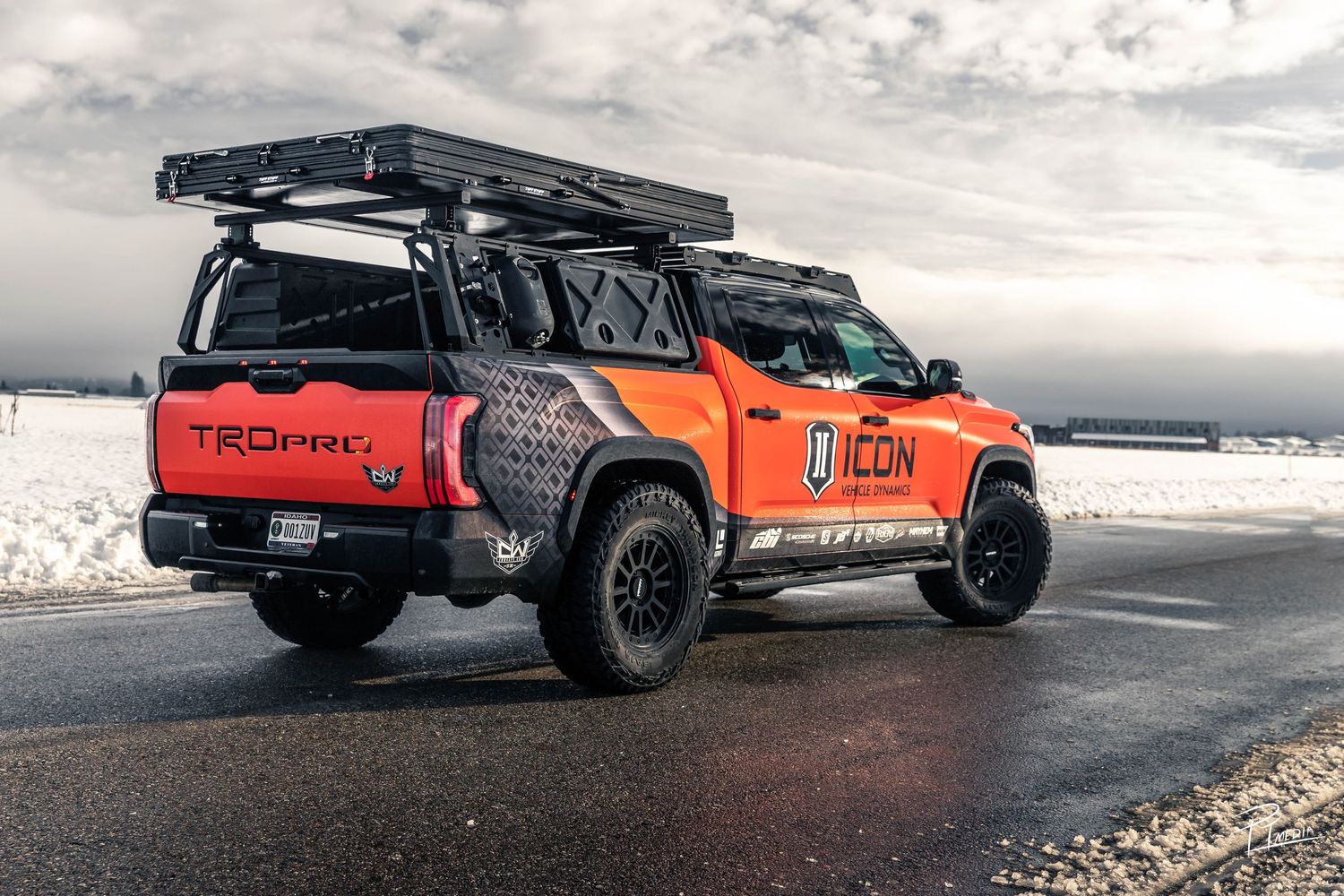 Custom orange Toyota Tacoma pickup truck with off-road modifications and roof rack parked on wet road at dusk.