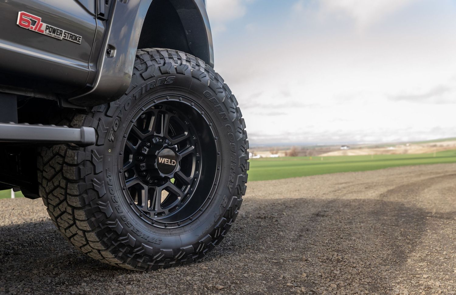 Closeup view of rugged off-road tires with aggressive tread patterns mounted on black wheels on a dirt path.