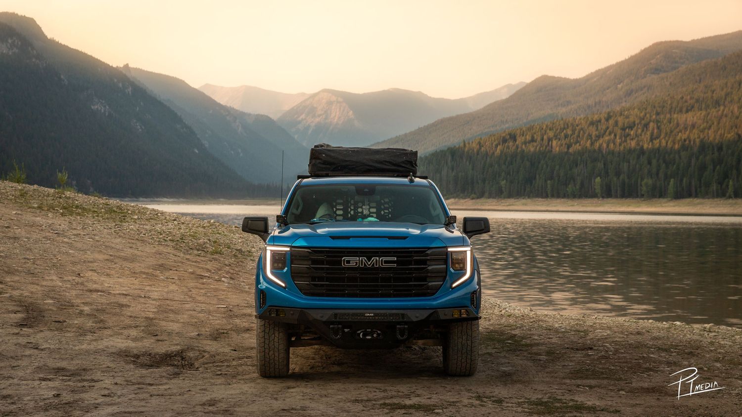 A panoramic view of a blue sports car parked along a scenic mountain lake at sunset.