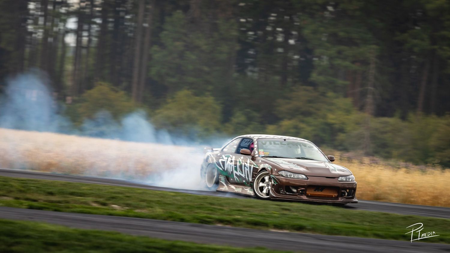 A red sports car drifts sideways on a racetrack creating smoke from tire burnout during a drift competition.