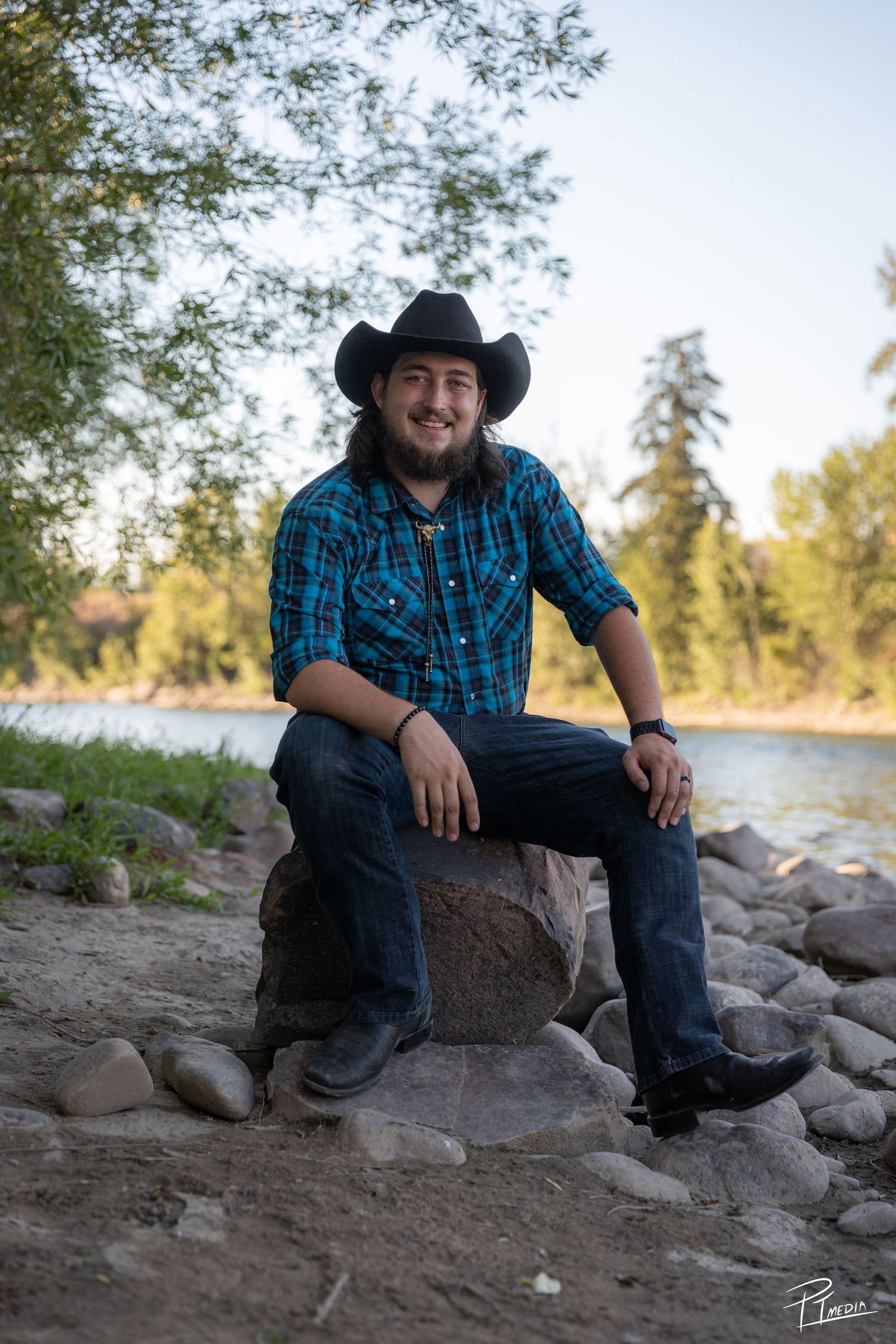 A person in western attire sits on rocky riverbank during golden hour wearing a cowboy hat and plaid shirt.